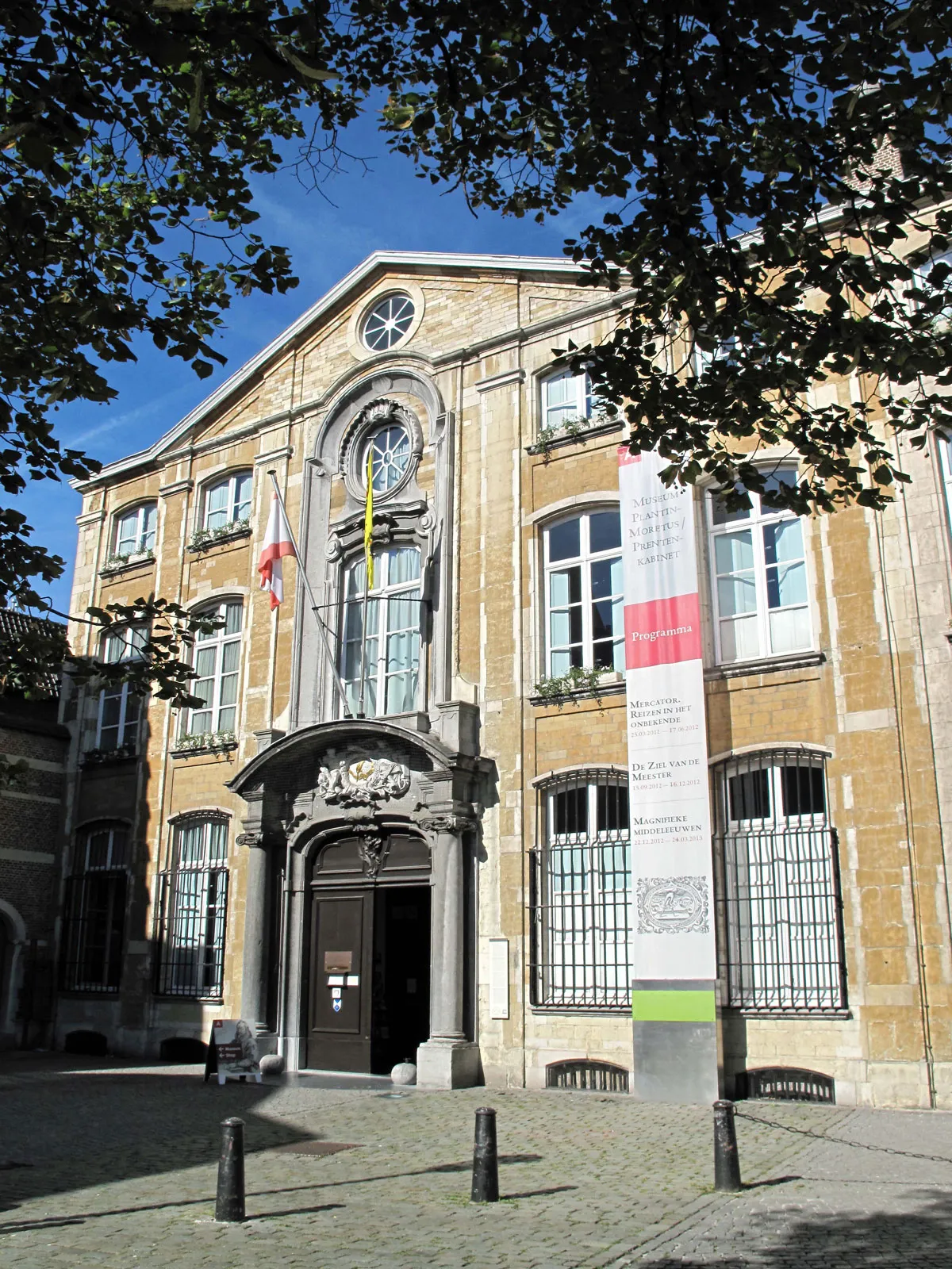 Fachada de piedra del Museum Plantin-Moretus con una ornamentada entrada arqueada, un rosetón central, banderas y una pancarta vertical, enmarcada por ramas de árboles contra un cielo azul