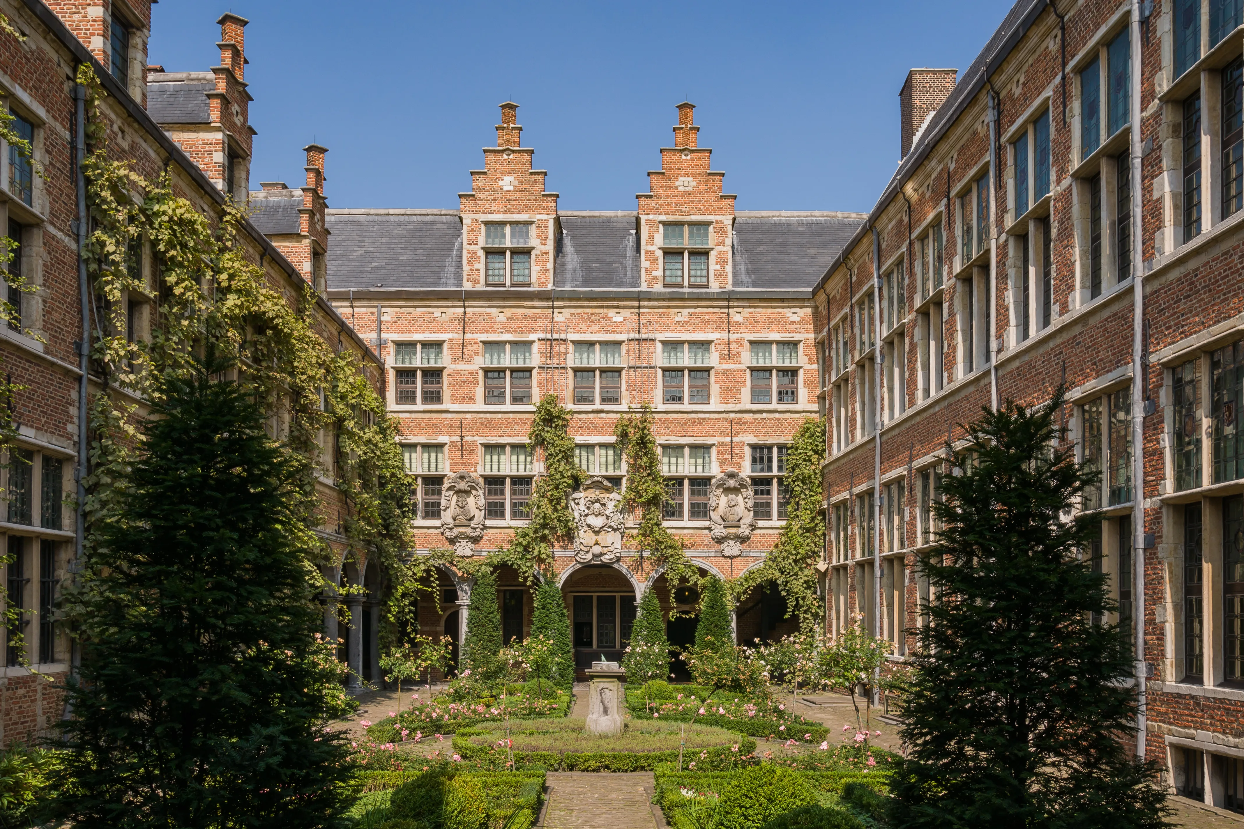 Jardín del patio del Museo Plantin-Moretus con edificios de ladrillo con gabletes escalonados, paredes cubiertas de hiedra y un jardín formal con setos recortados y árboles bajo un cielo azul.