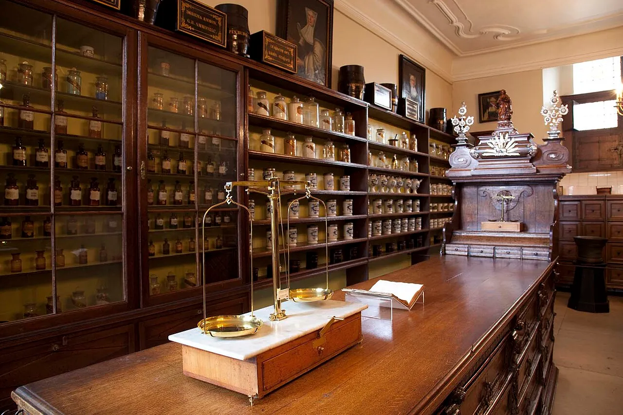 Historic pharmacy interior with tall dark wooden cabinets filled with glass jars and bottles, a long wooden counter with a brass balance scale, and an ornate wooden apothecary cabinet in the background