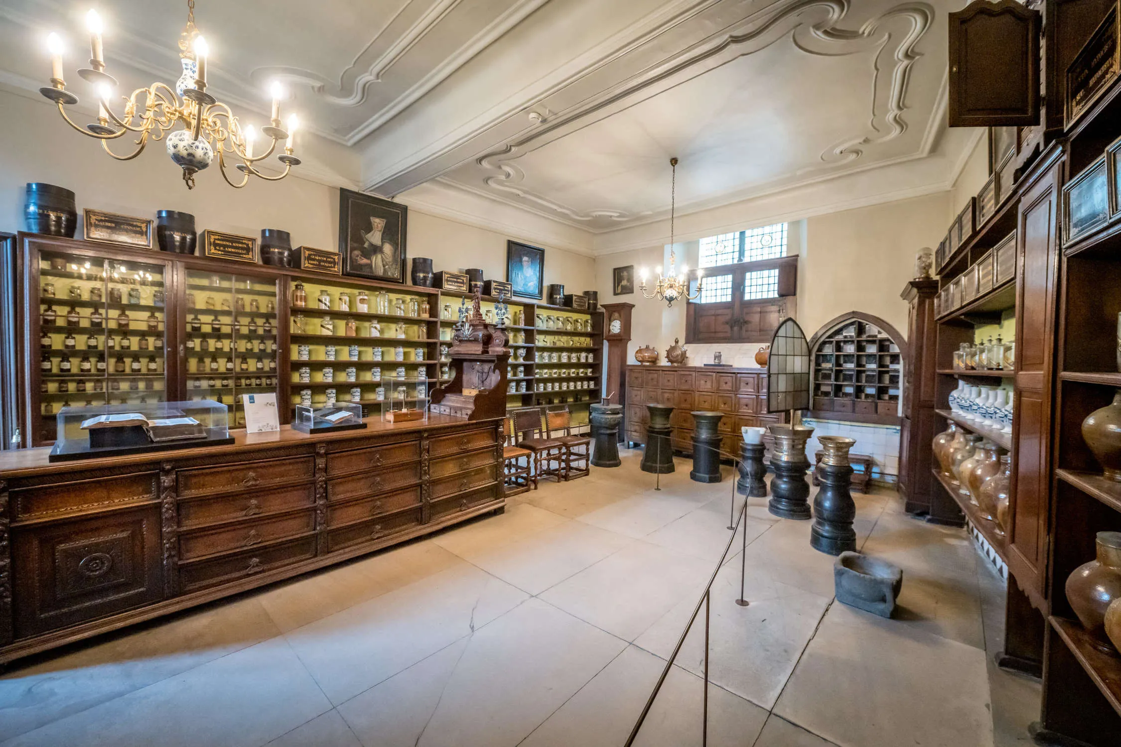 Historic pharmacy interior with tall wooden cabinets filled with rows of ceramic jars, an ornate chandelier, decorative ceiling moldings, and large wooden barrels on a stone floor at Museum Sint-Janshospitaal