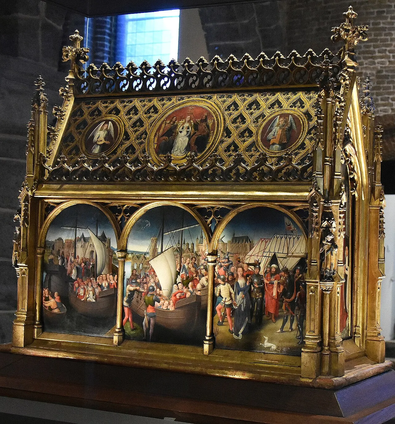 Ornate gilded Gothic reliquary shrine with painted narrative panels depicting medieval scenes of ships and figures, displayed in a museum setting