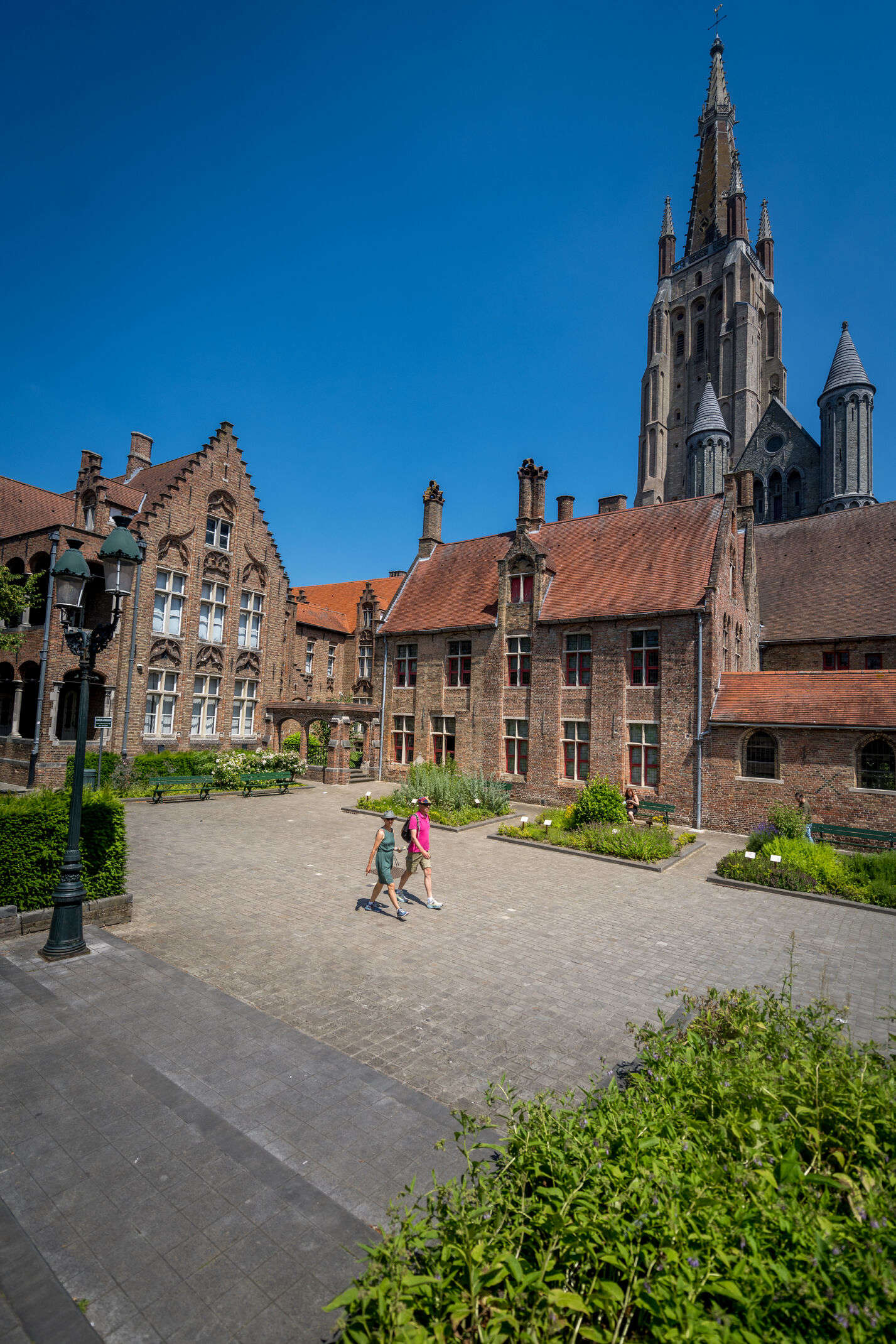Courtyard of Museum Sint-Janshospitaal with historic brick buildings and a Gothic church spire rising in the background under a clear blue sky
