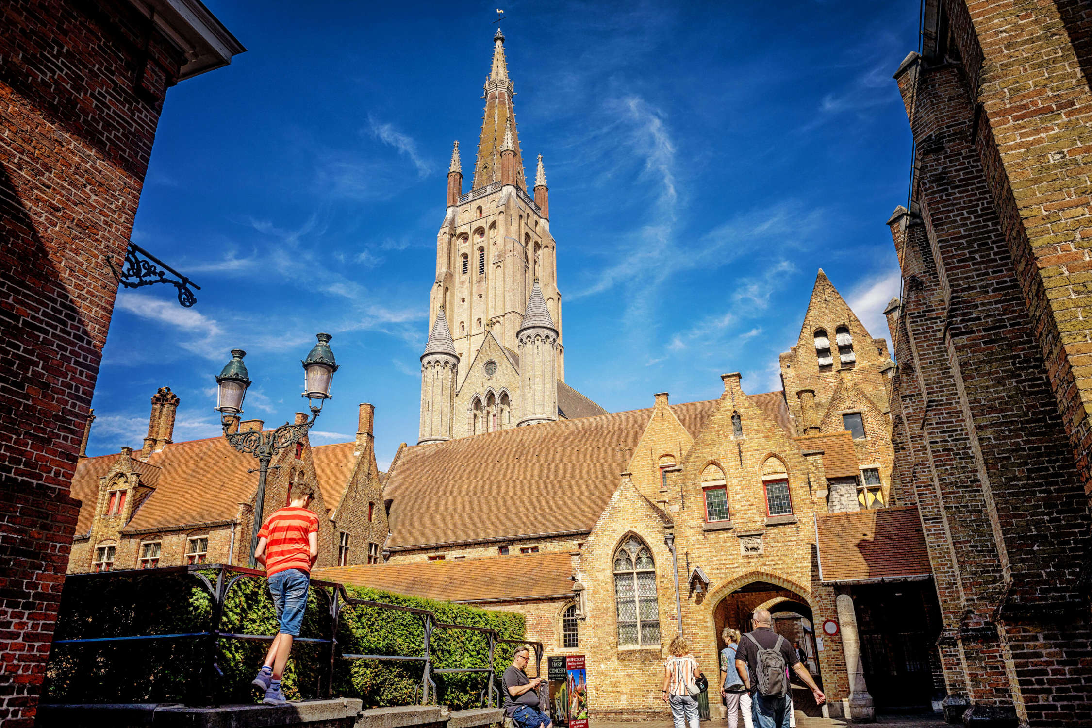 Exterior view of the Sint-Janshospitaal courtyard with brick buildings, an arched gateway, ornate lamppost, and the tall Gothic spire of the Church of Our Lady rising behind against a bright blue sky, with visitors walking through the scene.