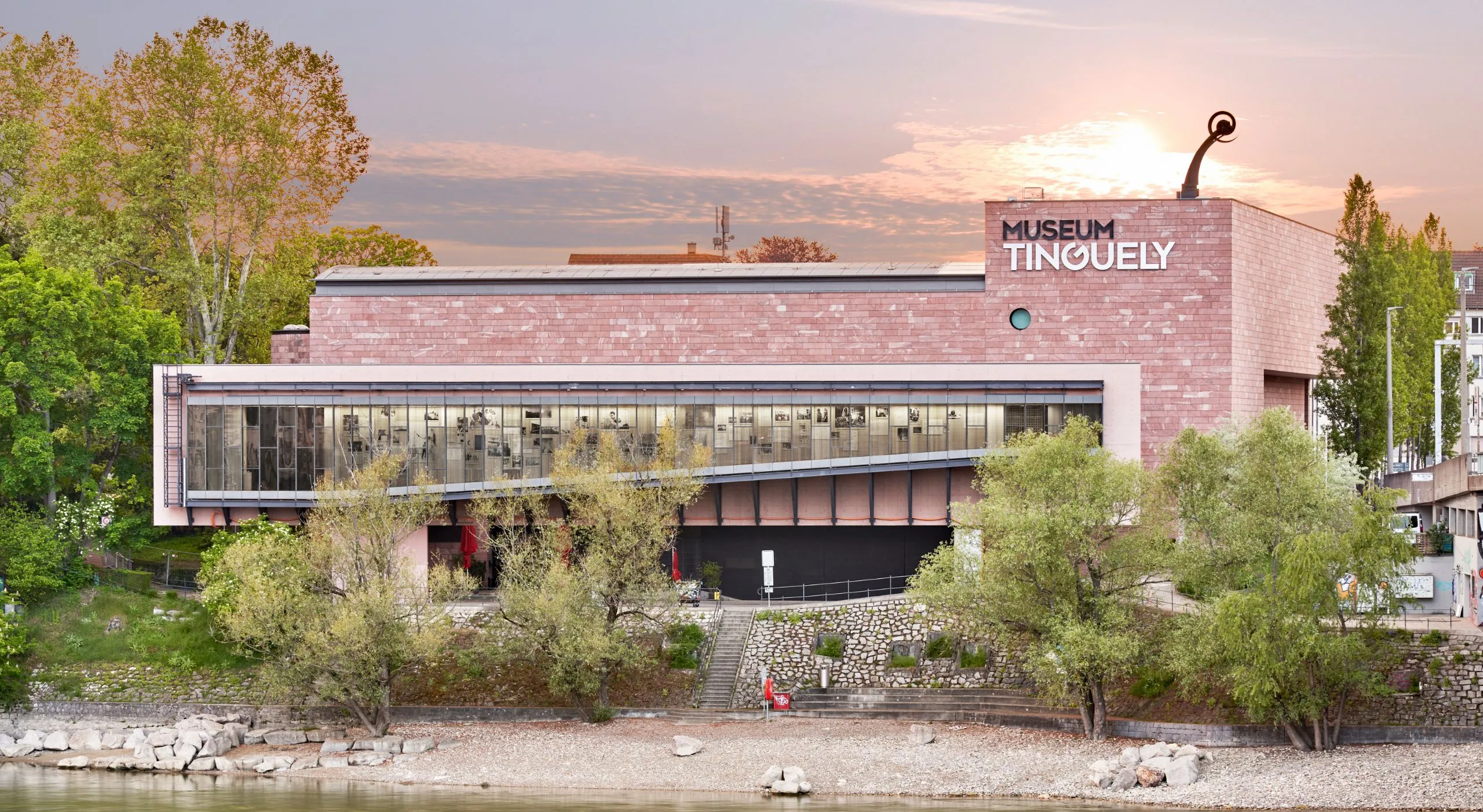 Widok z zewnątrz na Museum Tinguely, budynek z różowej cegły z dużymi szklanymi oknami i nazwą muzeum na górnej fasadzie, zwieńczony rzeźbą kinetyczną, otoczony drzewami przy rzece o zachodzie słońca