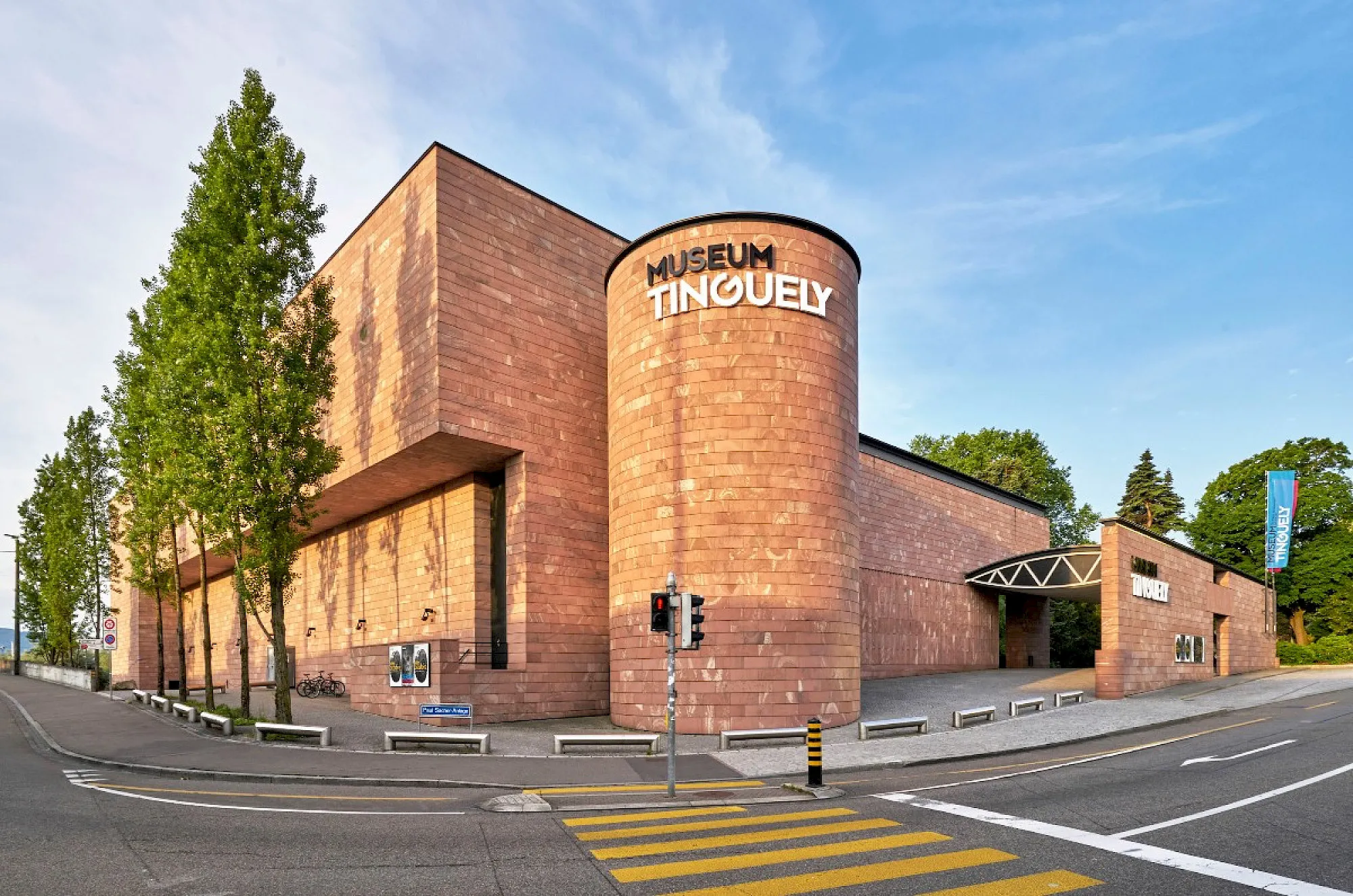 Widok na zewnątrz Museum Tinguely przedstawiający czerwoną, ceglaną, cylindryczną wieżę z białym napisem, otoczoną zielonymi drzewami i ulicą z przejściem dla pieszych pod błękitnym niebem.