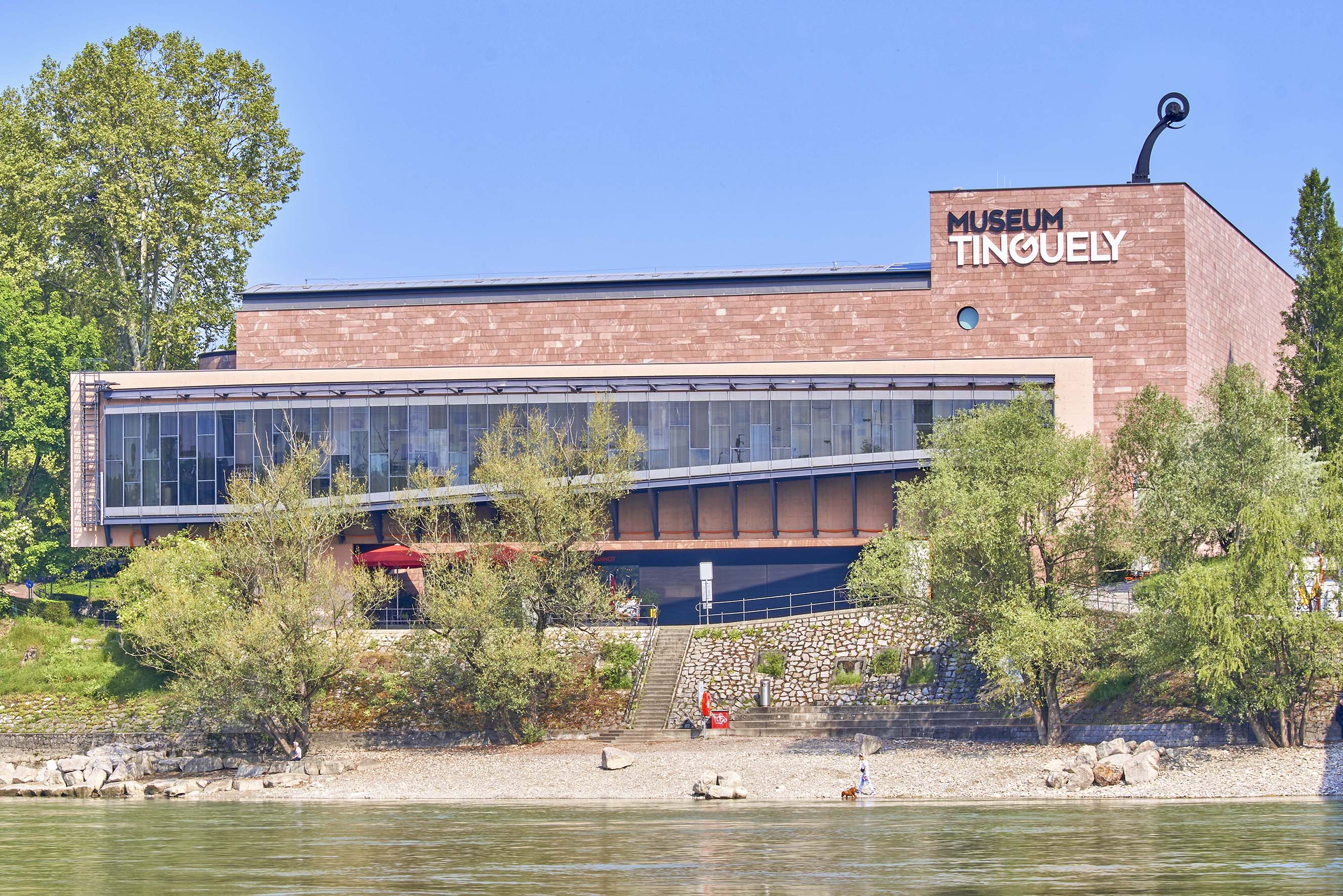 Ceglany budynek Museum Tinguely widziany z drugiego brzegu Renu, z drzewami, małą plażą i nazwą muzeum widoczną na fasadzie.