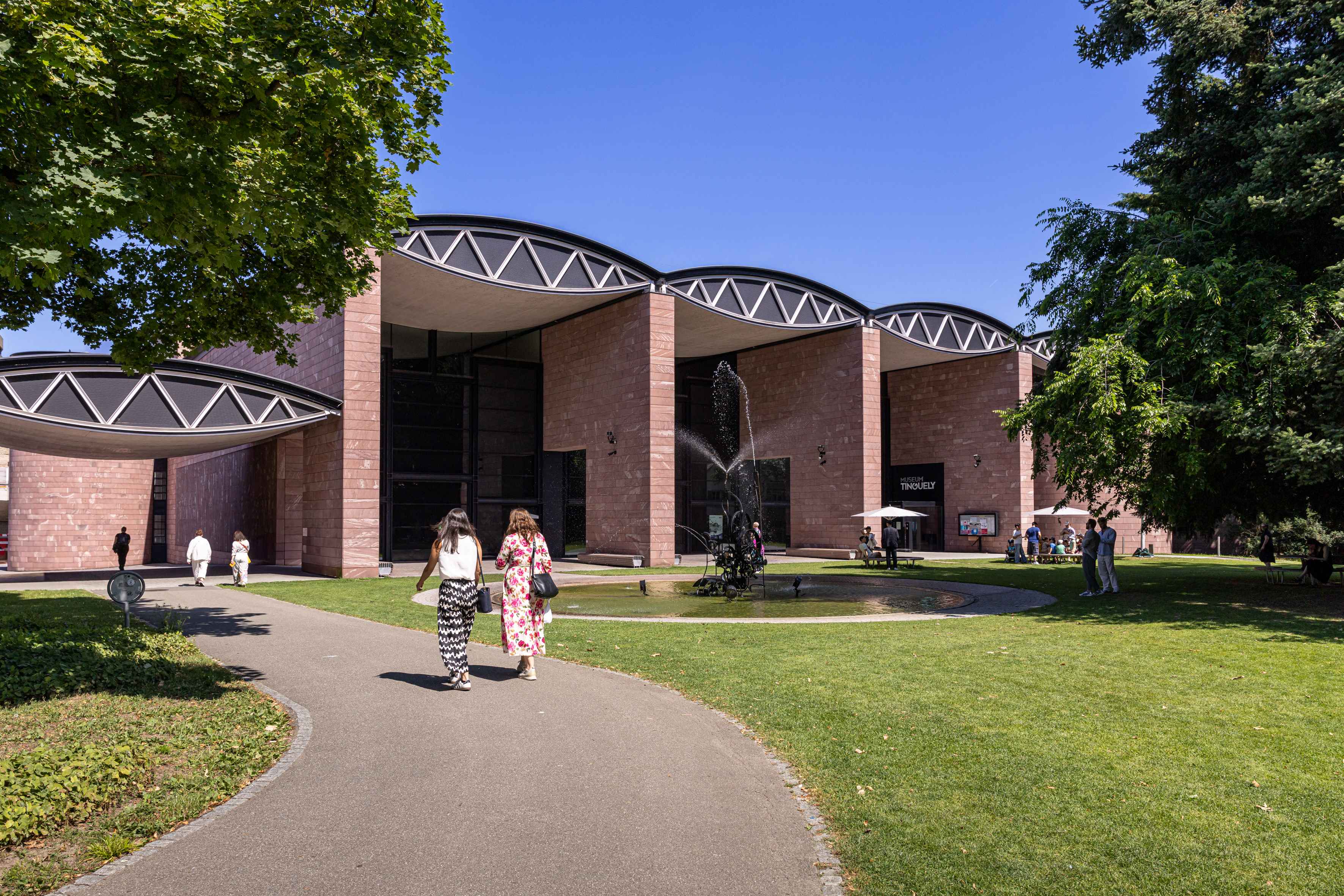 Widok na zewnątrz Museum Tinguely z charakterystyczną zakrzywioną fasadą z cegły i falistą linią dachu, otoczony zielonymi trawnikami i drzewami, ze zwiedzającymi spacerującymi ścieżką