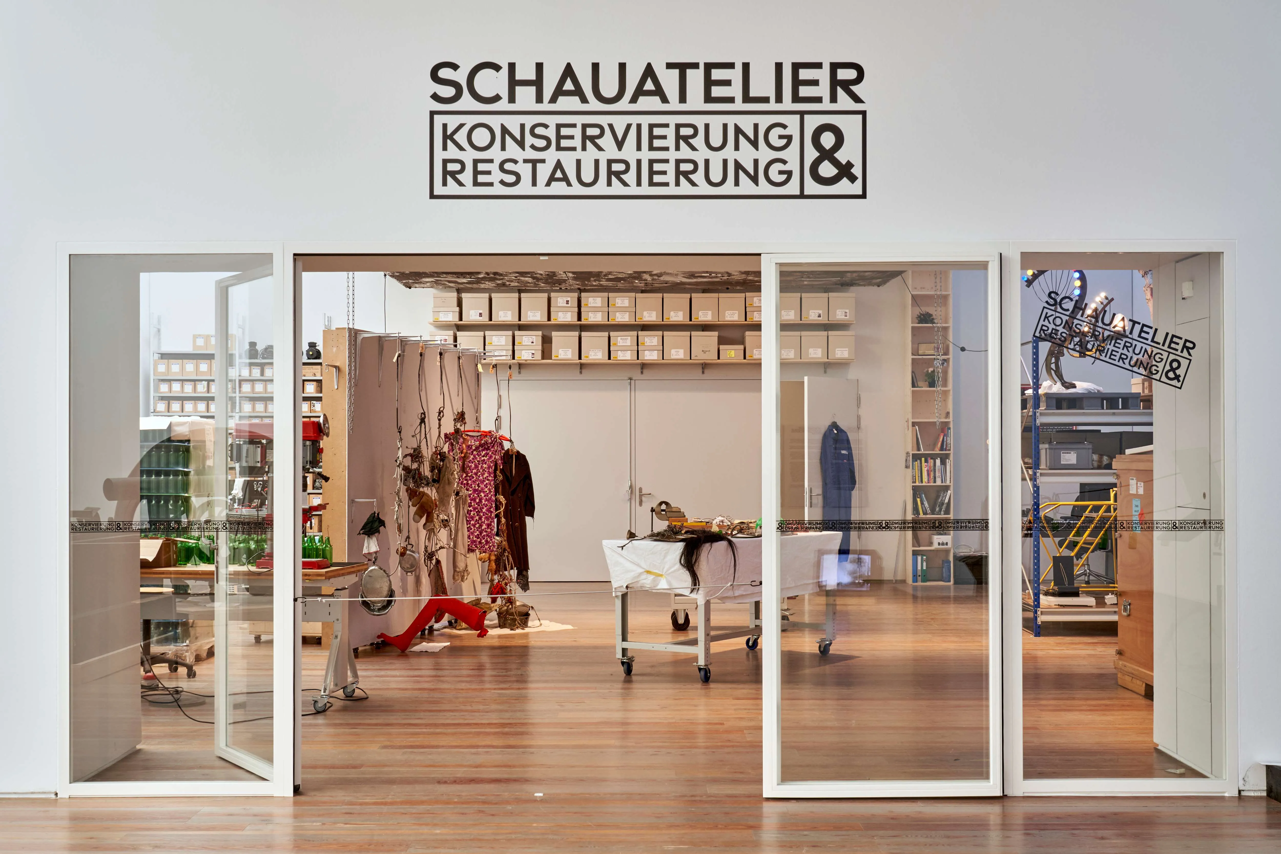 Widok wnętrza pracowni konserwatorsko-restauratorskiej Schauatelier w Museum Tinguely, ze szklanymi drzwiami, drewnianymi podłogami i dziełami sztuki poddawanymi renowacji.
