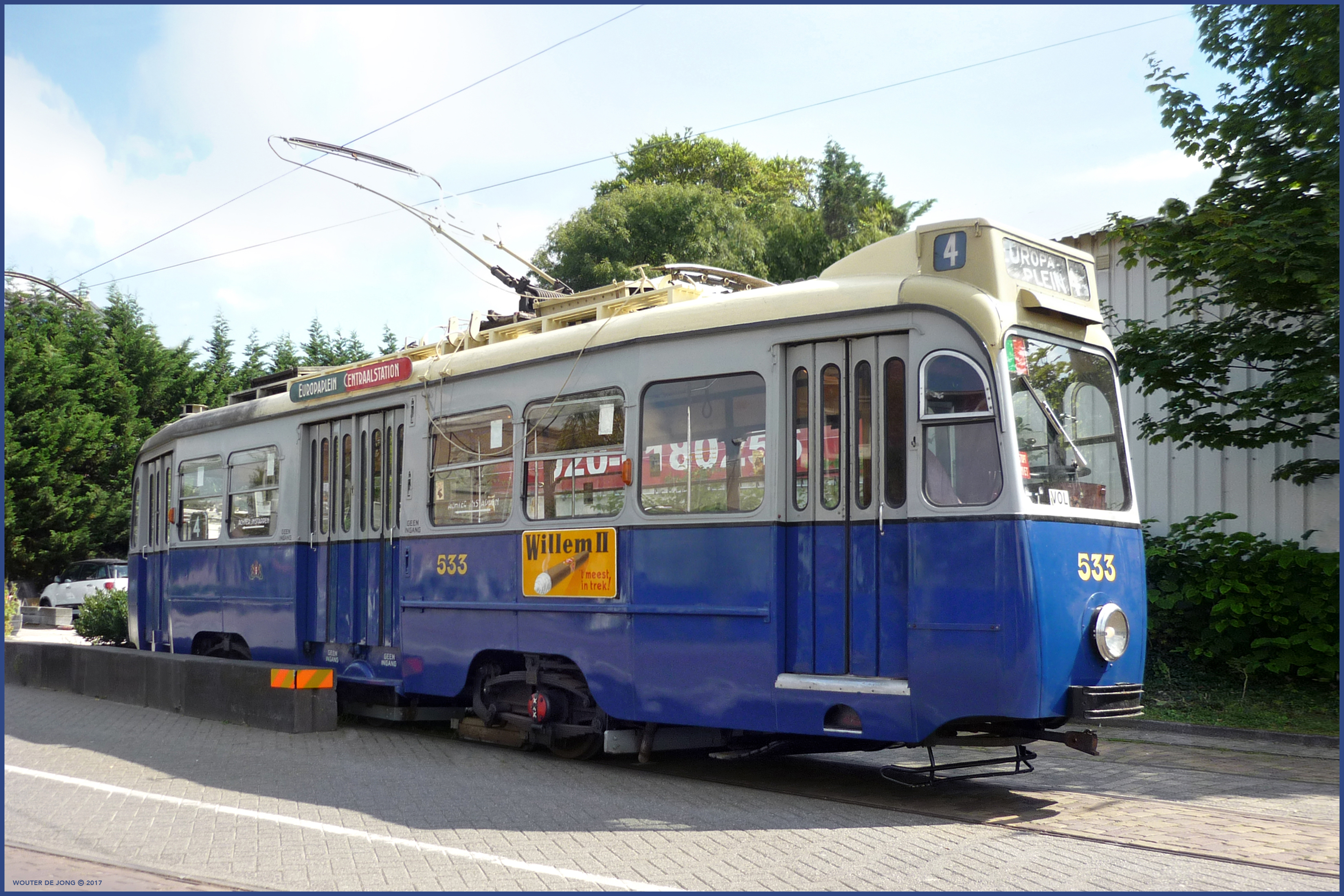 Niebieski i kremowy zabytkowy tramwaj numer 533 na torach z przewodami napowietrznymi i drzewami w tle
