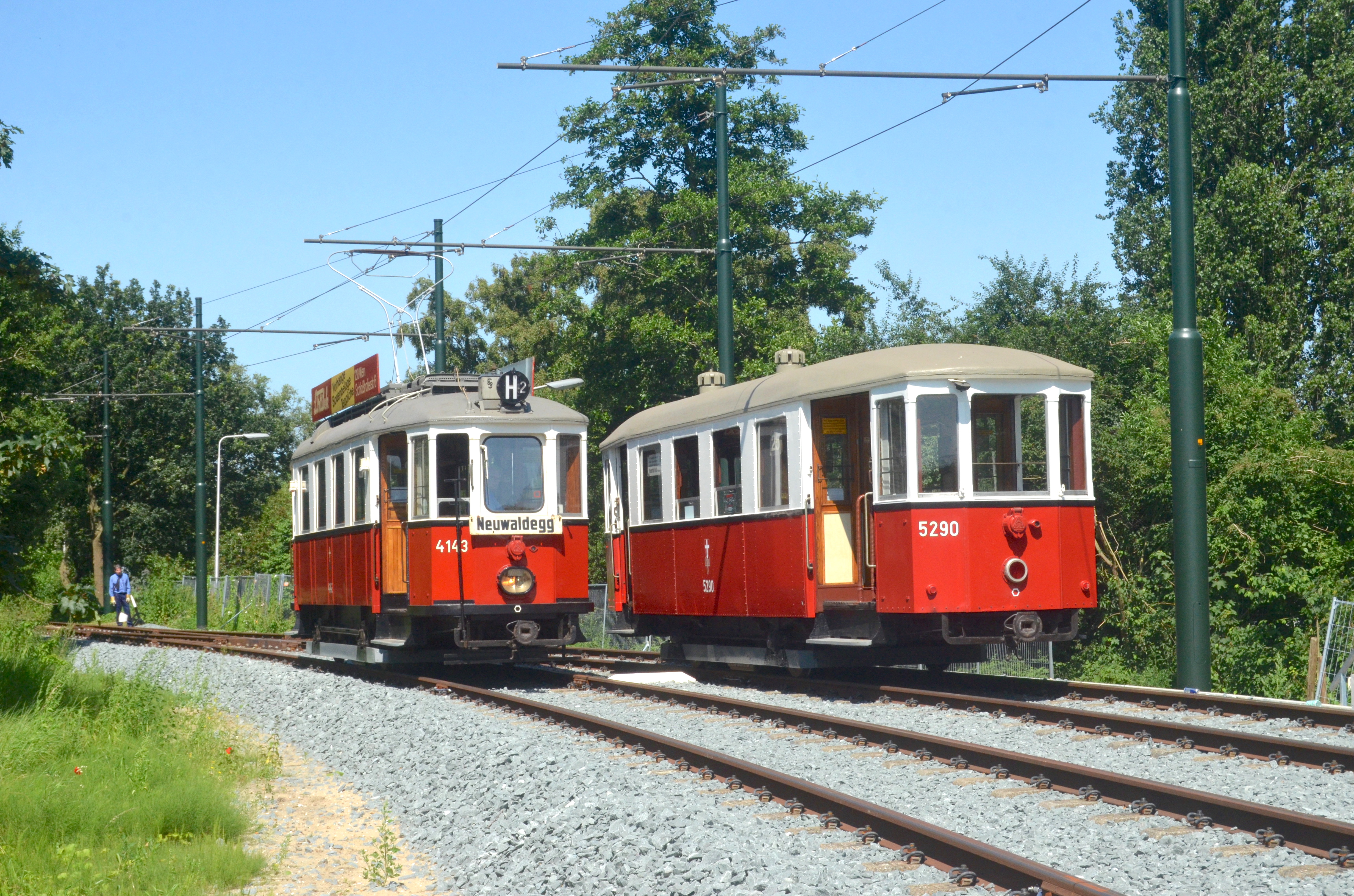 Dwa zabytkowe tramwaje w kolorach czerwonym i kremowym na torach kolejowych pod liniami napowietrznymi, z drzewami i czystym błękitnym niebem
