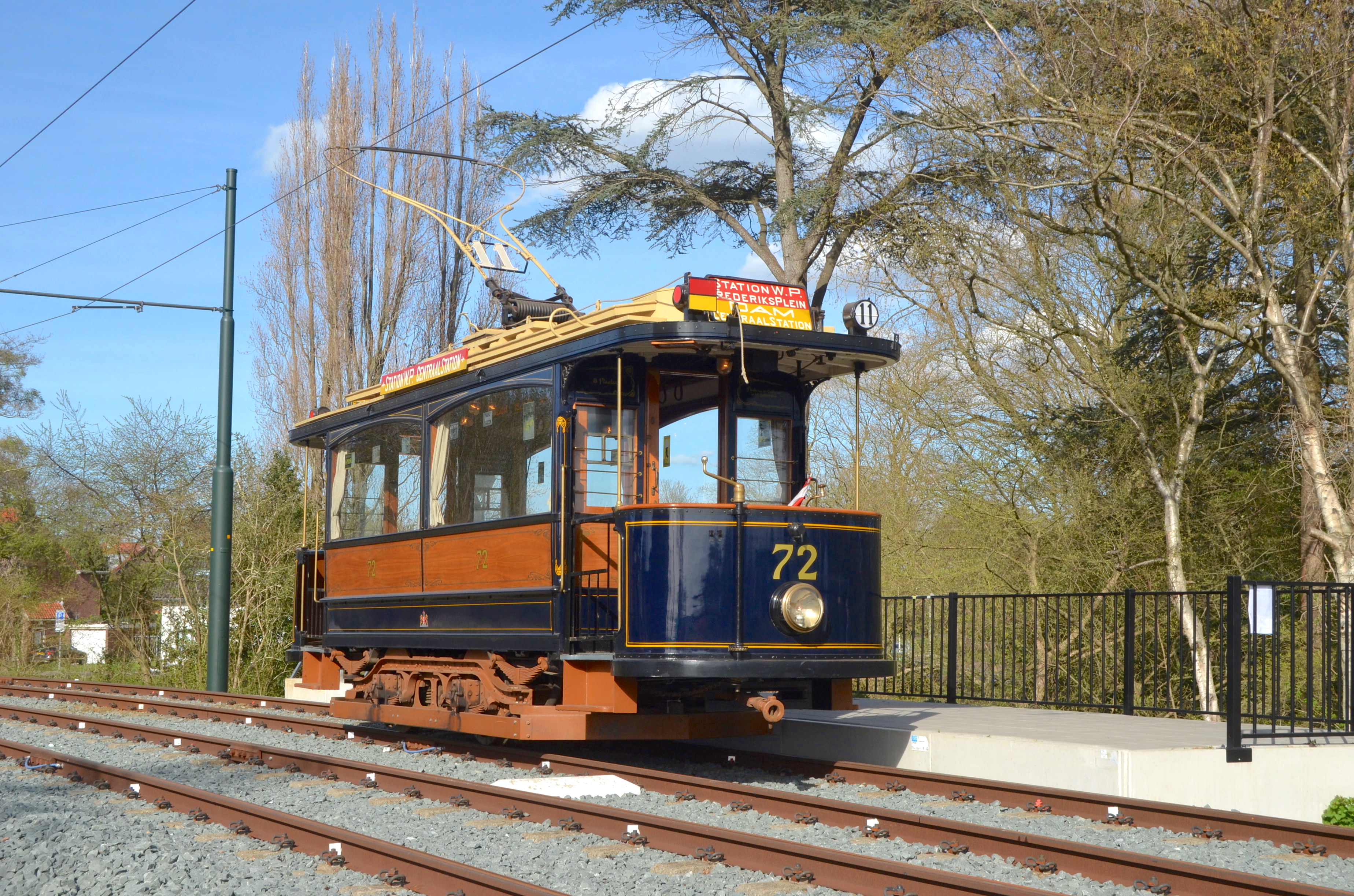 Niebiesko-pomarańczowy zabytkowy tramwaj numer 72 na torach z przewodami napowietrznymi i drzewami w tle