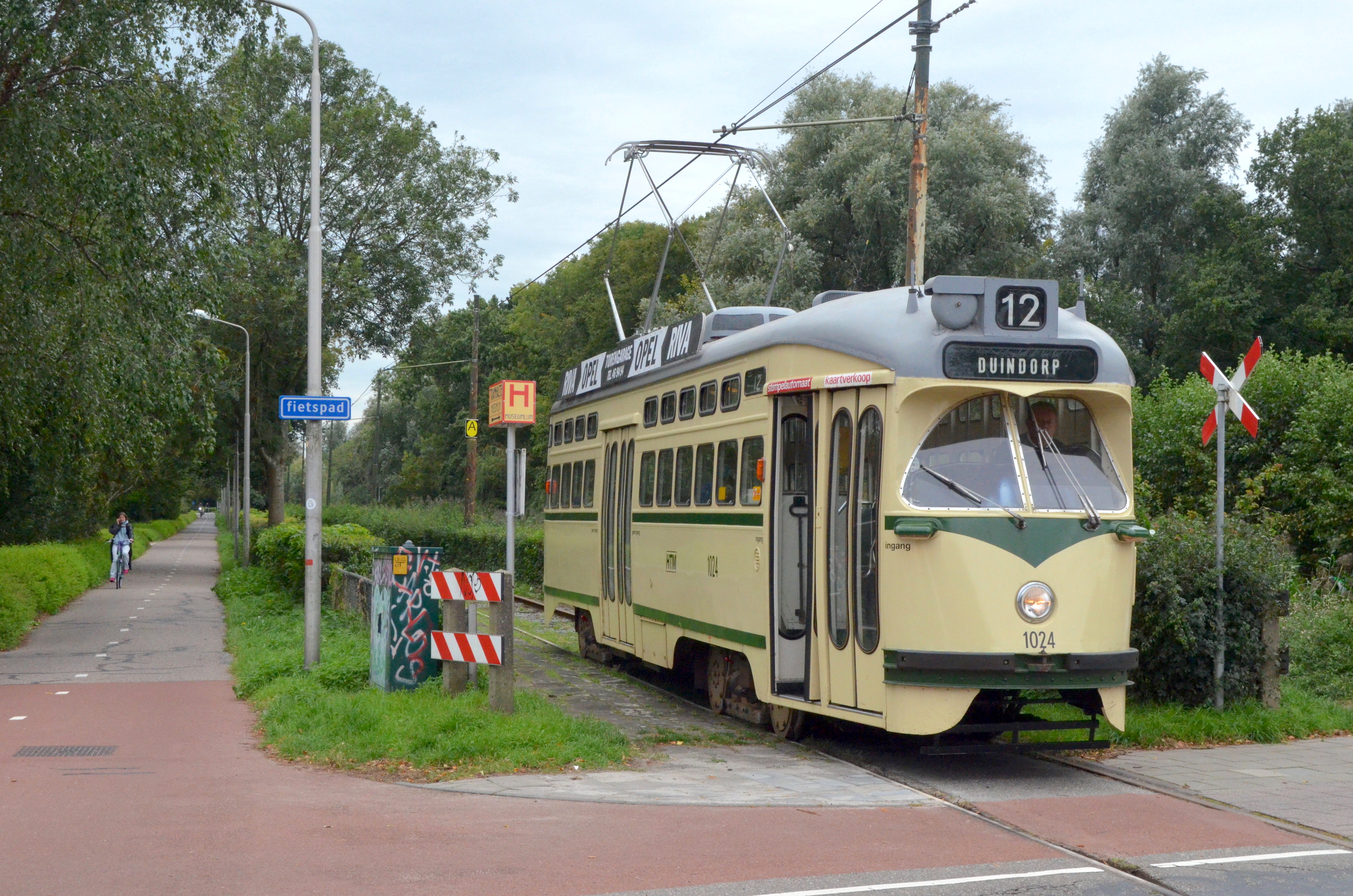 A cream and green vintage tram with the number 12 on its destination sign, stopped at a track junction with overhead wires and a red and white crossing signal.