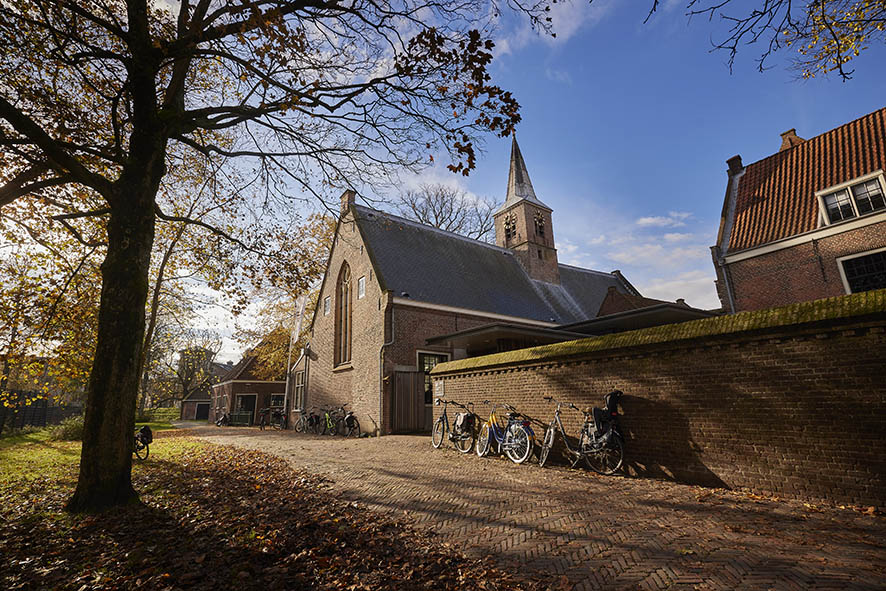 Bakstenen kerk-stijl gebouw met een spits, rode pannendaken en een stenen muur. Fietsen leunen tegen de muur, omringd door herfstbomen en gevallen bladeren onder een helderblauwe hemel.