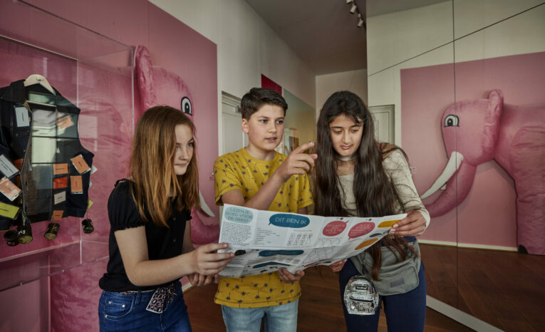 Drie kinderen bekijken samen een museumkaart in een kamer met roze muren, een olifantenmuurschildering en tentoonstellingen.