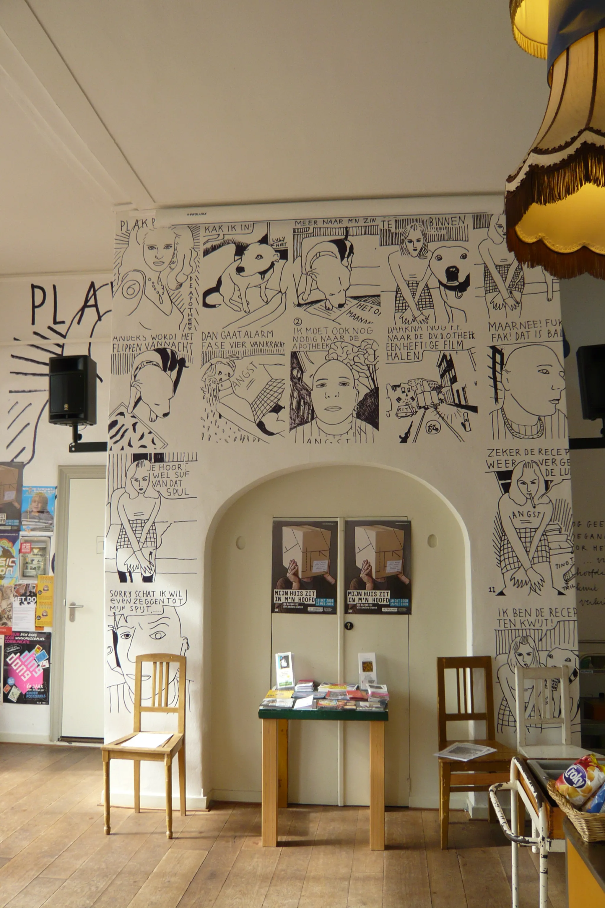 Binnenmuur bedekt met zwart-wit illustraties in stripstijl en tekstpanelen, met houten stoelen en een tafel ervoor.