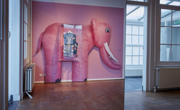 Roze olifantvormige structuur met een glazen vitrine erin, tegen een roze muur in een museumzaal met houten vloeren en grote ramen