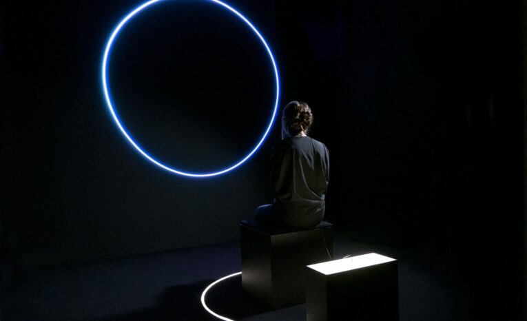 Una persona se sienta frente a una instalación de círculo de neón azul brillante en una habitación oscura, con un pequeño pedestal iluminado cerca.