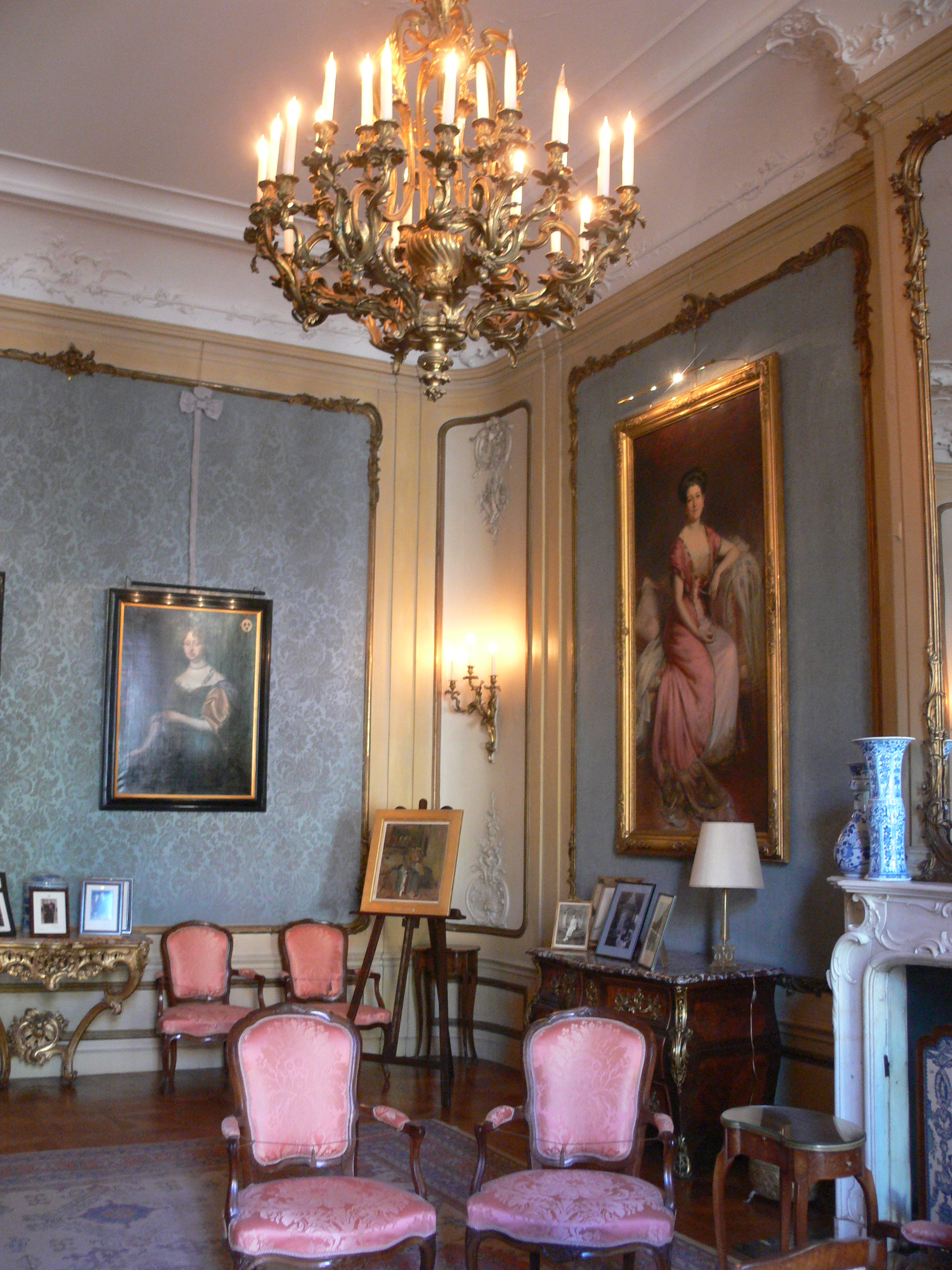 Ornate room with a large chandelier, pink chairs, and framed portraits on patterned walls