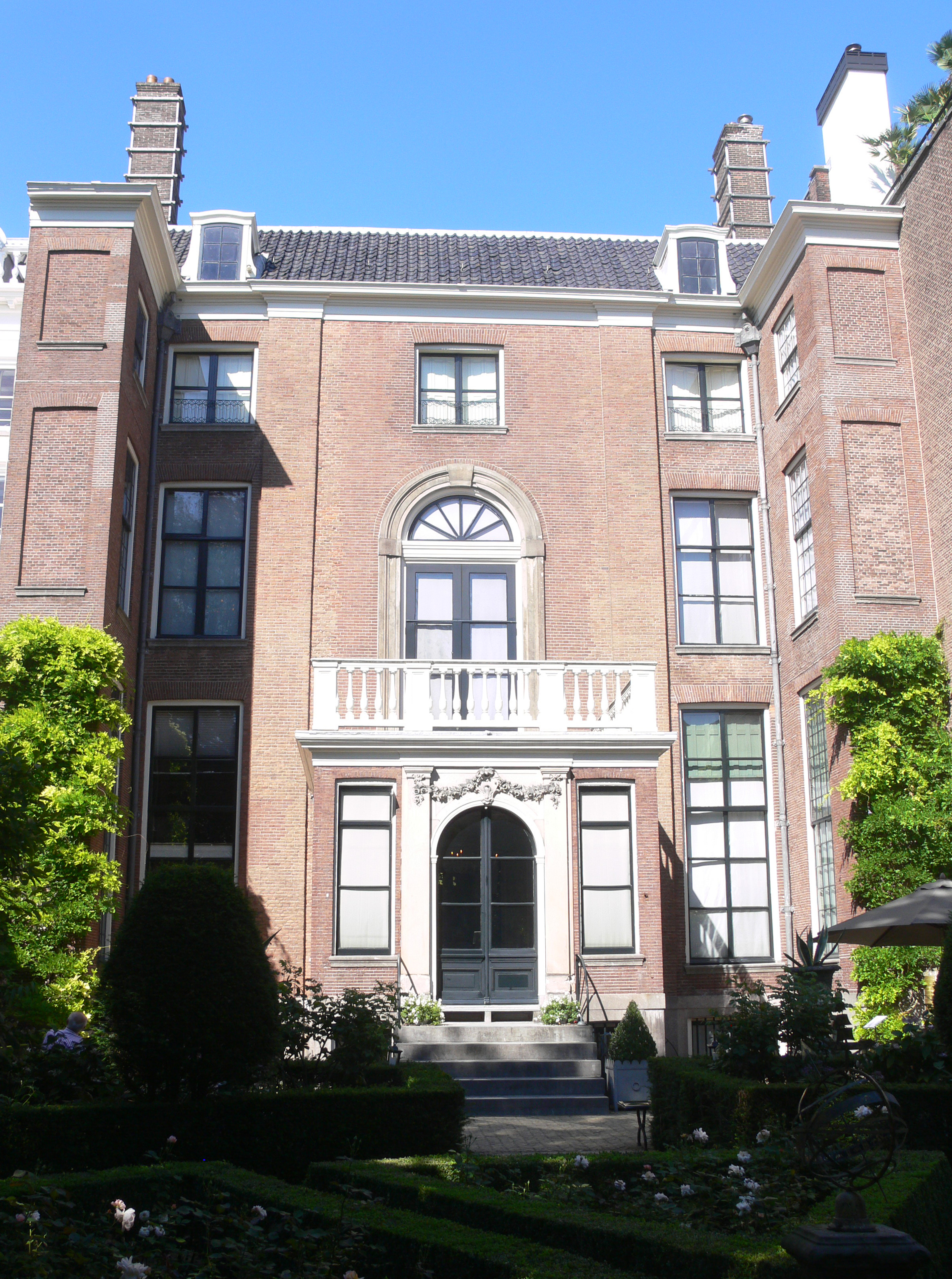 Historic brick canal house with arched windows, central balcony, and manicured garden under clear blue sky