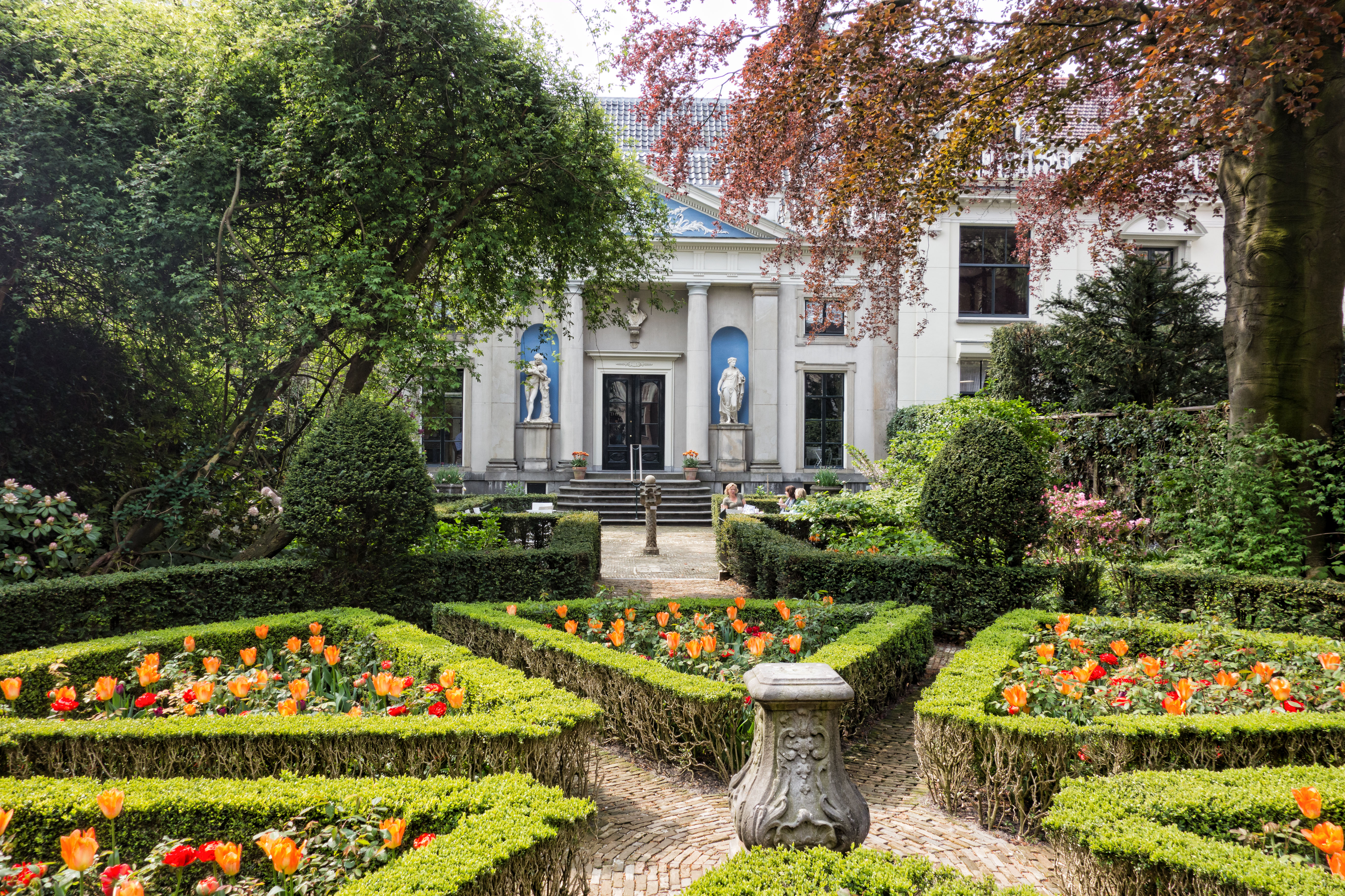 Formal garden with manicured hedges and orange flowers leading to the white facade of Museum Van Loon with columns and statues