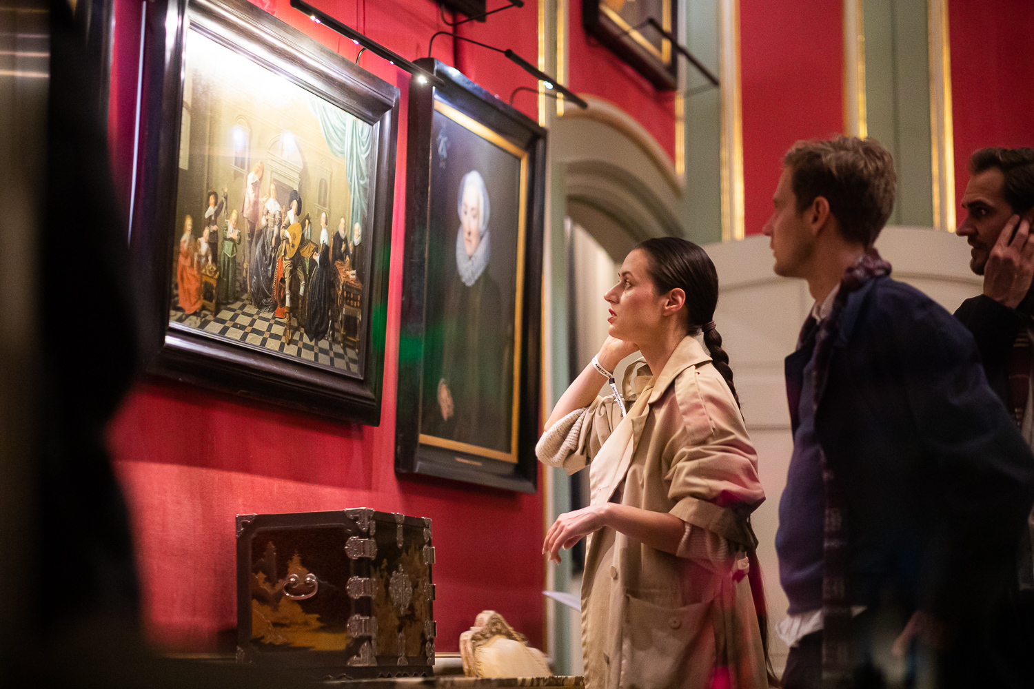 People viewing framed historical paintings on red walls inside a museum room with a decorative cabinet display