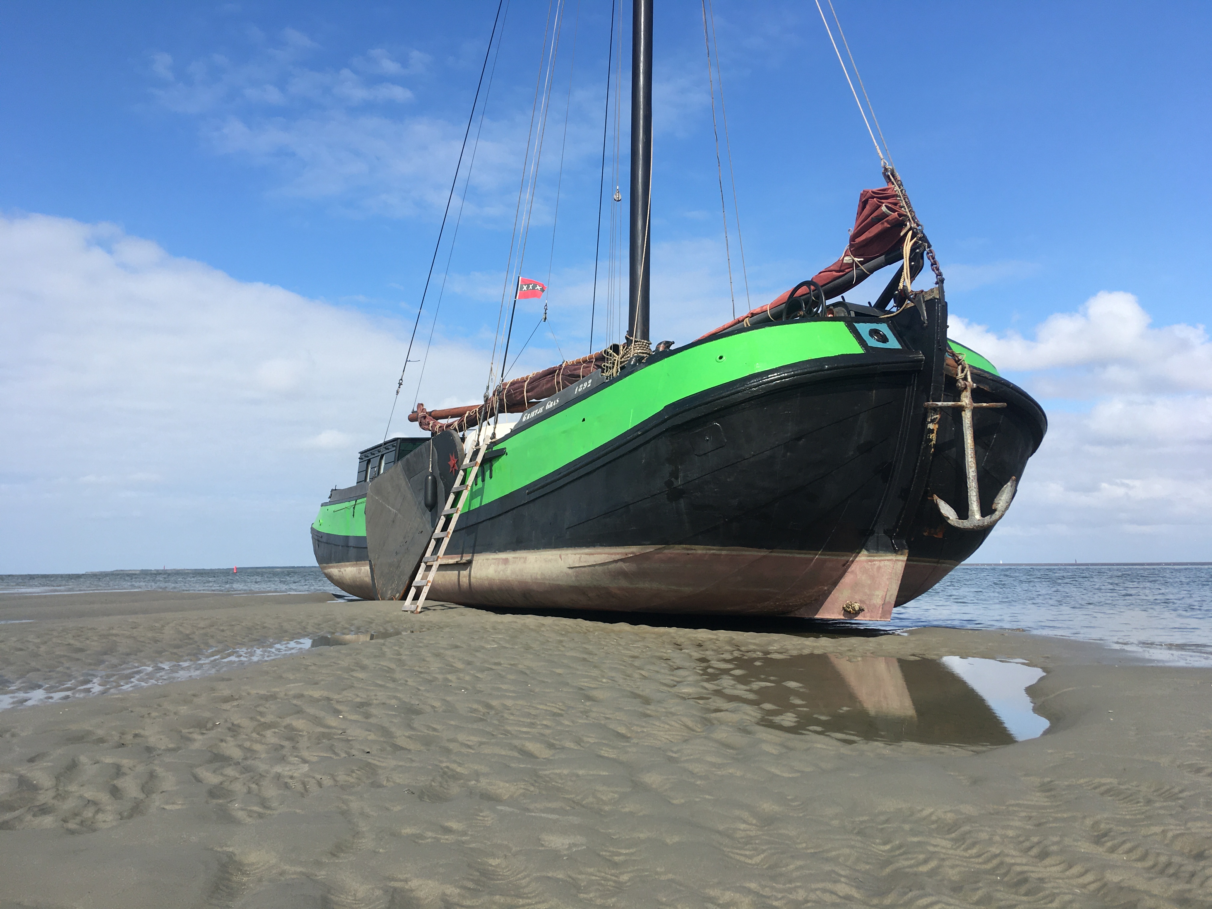 Barco de vela histórico verde y negro descansando en la orilla arenosa con cielo azul y nubes dispersas