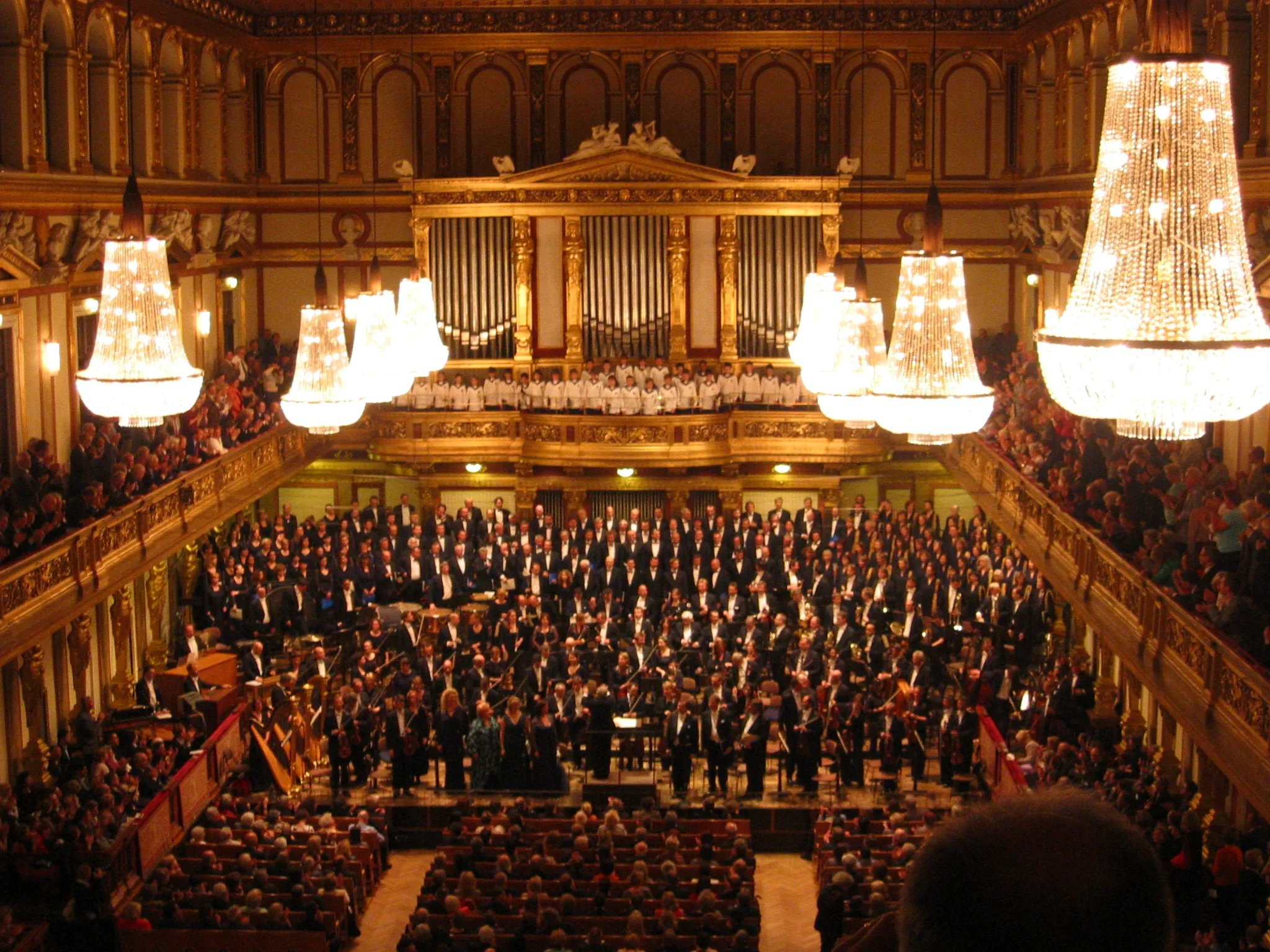 Wnętrze Złotej Sali Musikverein z wielką orkiestrą na scenie, ozdobnymi żyrandolami i pełną publicznością.
