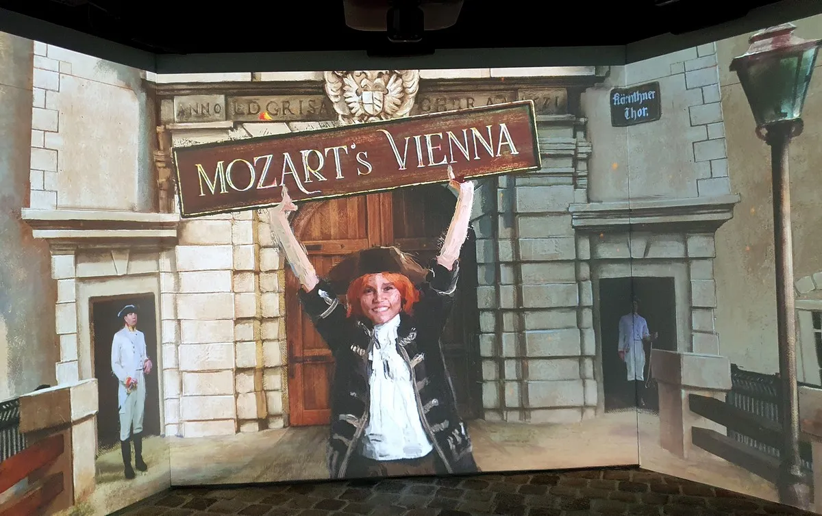 A person in historical clothing holding a sign that says 'MOZART'S VIENNA' in front of a painted building facade with a projection of a street scene.