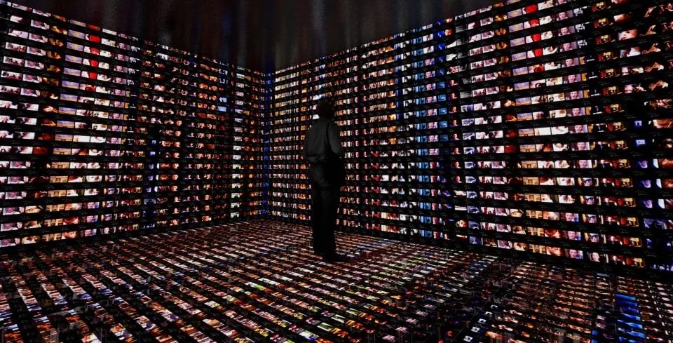 Person standing in a dark room surrounded by illuminated screens on walls and floor