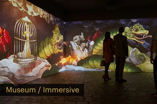 Dos personas de pie en una habitación oscura mirando una gran proyección de una escena surrealista y colorida con una jaula de pájaros, un dragón y llamas en las paredes.