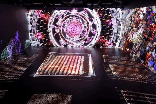 A dark exhibition space with a large circular projection on the back wall and a rectangular projection on the floor, surrounded by scattered light patterns.
