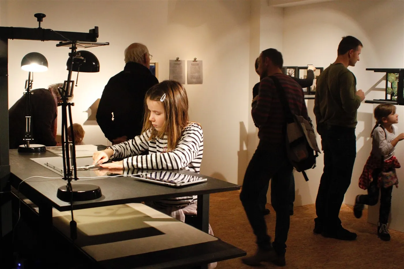A young girl in a striped shirt uses a light table with film strips while other visitors observe exhibits in a dimly lit museum gallery.