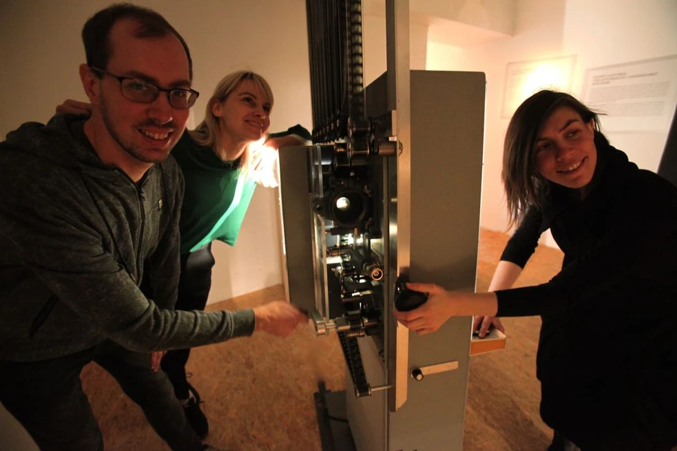 Three people smiling and operating a large film projector in a museum setting