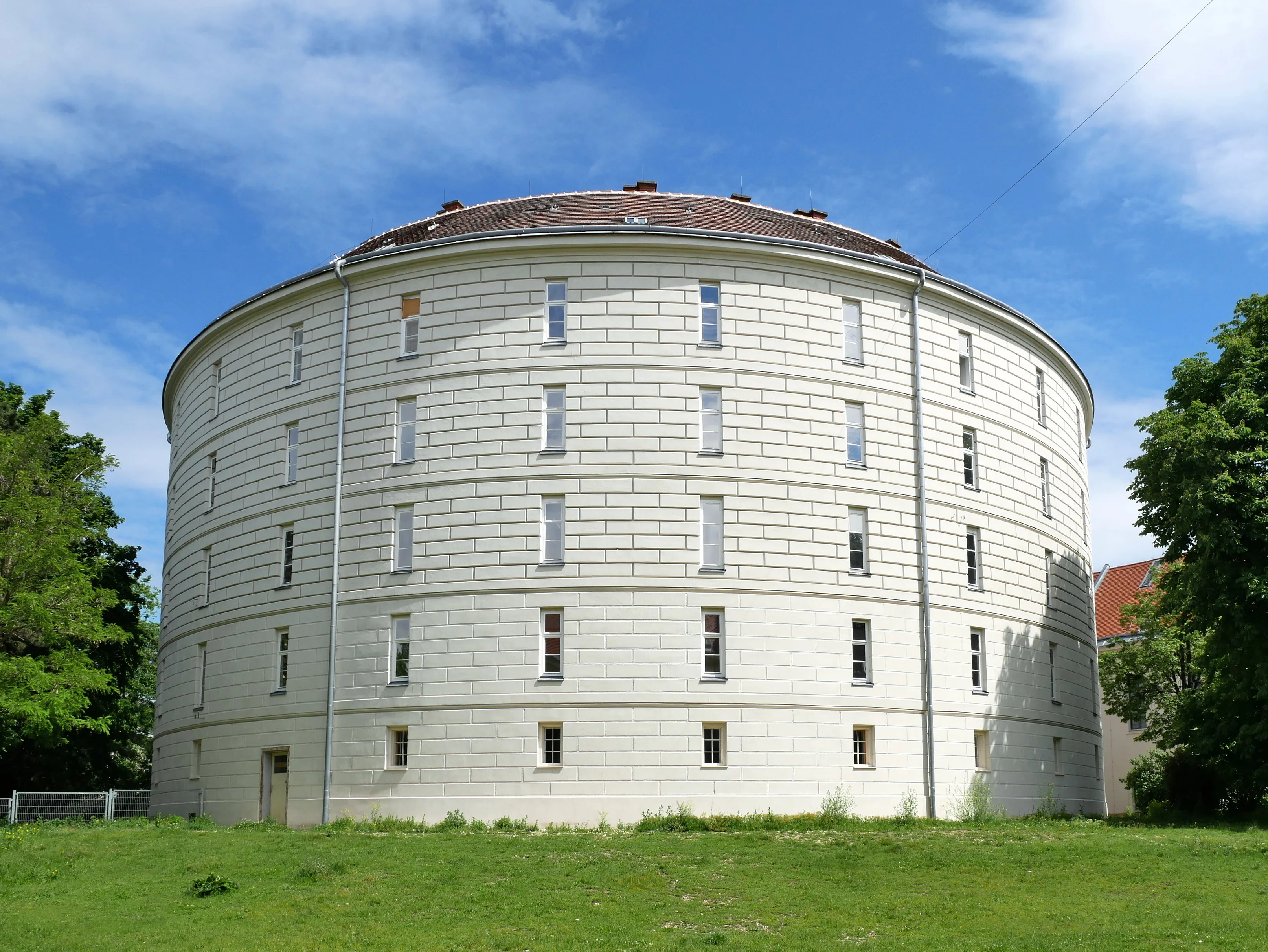 Circular white stone building with a domed roof and multiple windows, surrounded by green grass and trees under a blue sky