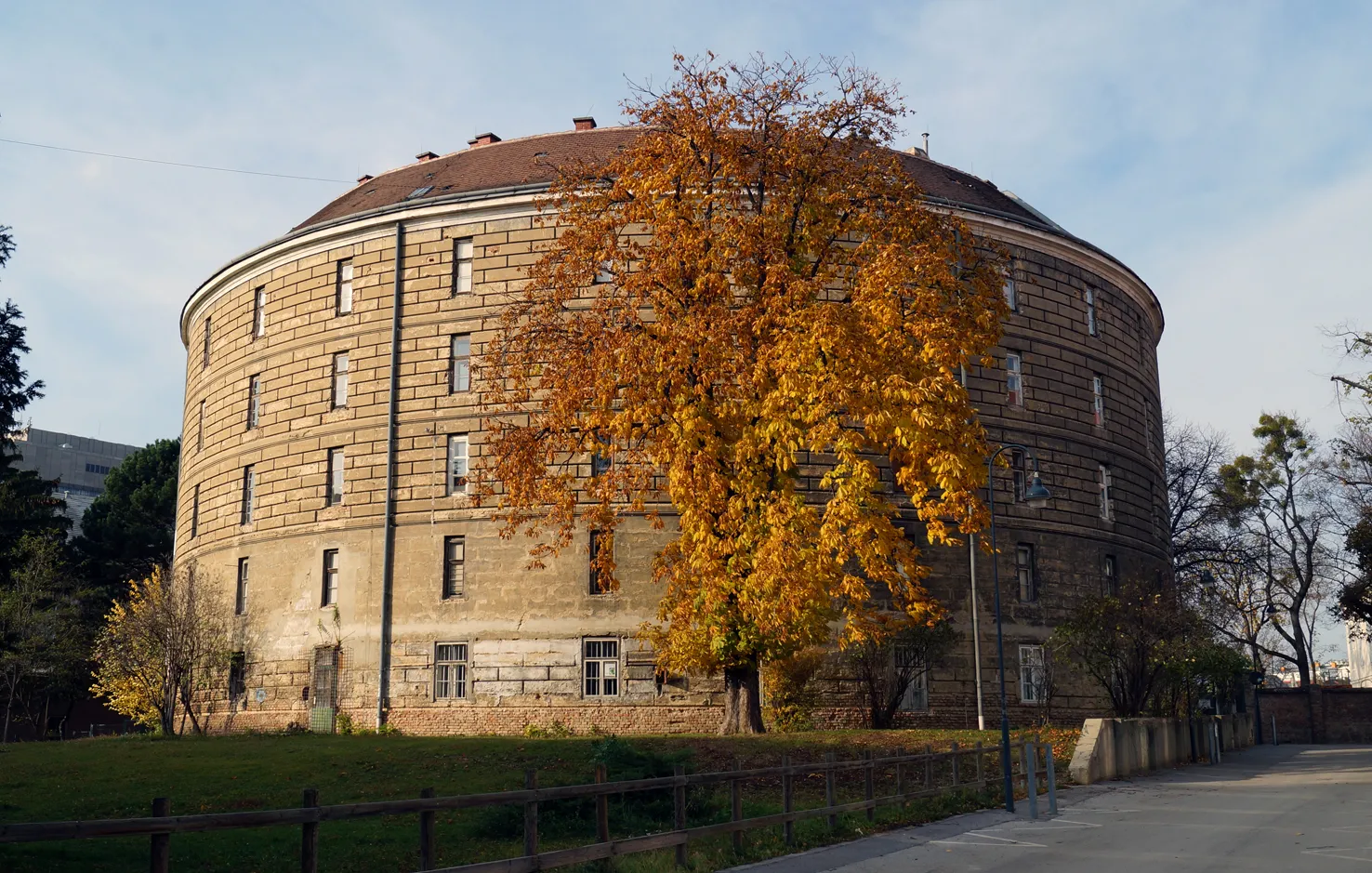 A large cylindrical stone building with a domed roof and many windows, surrounded by trees with autumn leaves