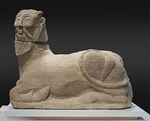 Stenen sfinxsculptuur met een menselijk hoofd en een leeuwenlichaam op een wit voetstuk