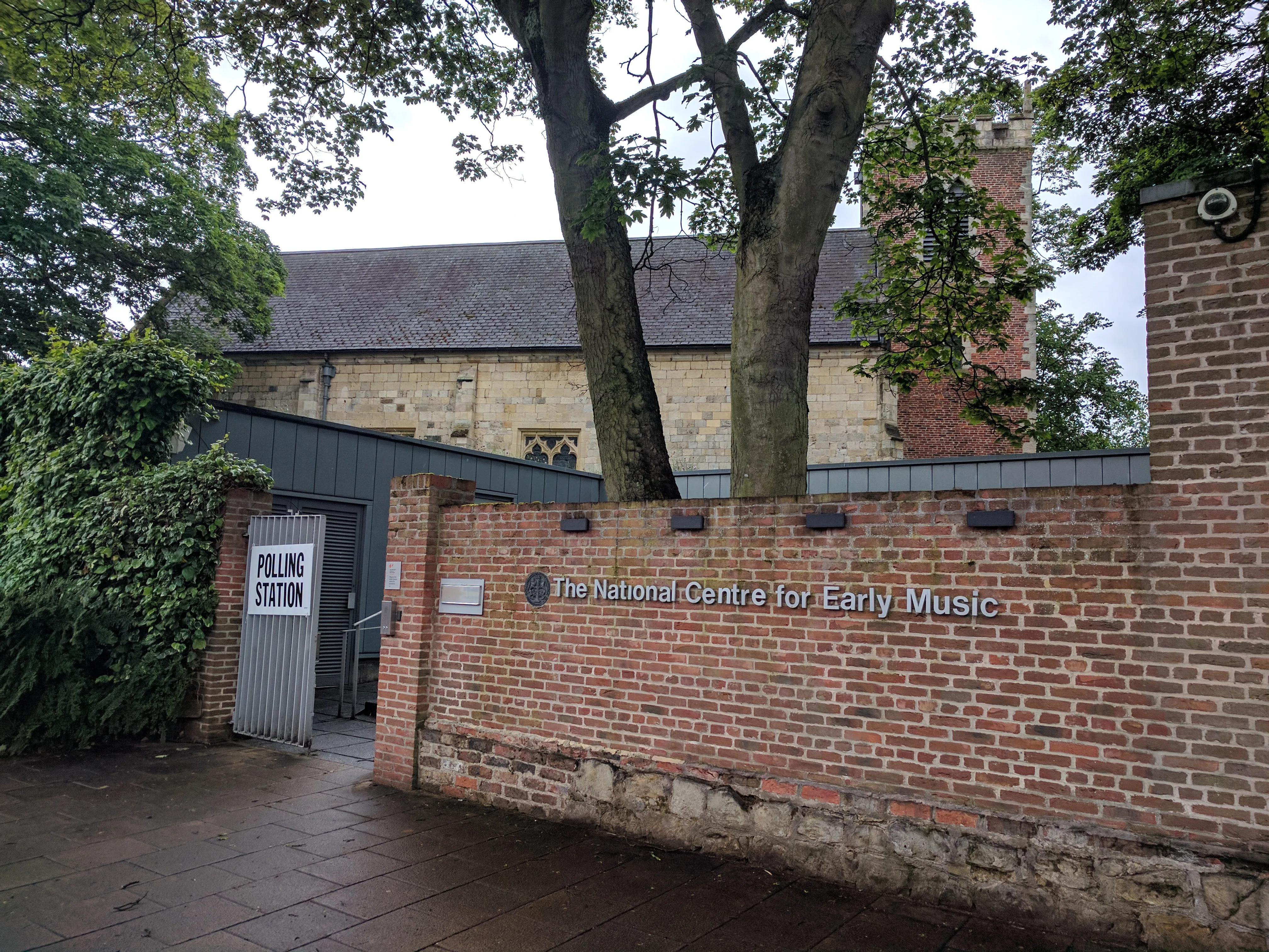Ceglany mur z szyldem 'The National Centre for Early Music' i bramą z oznaczeniem 'Polling Station', a w tle, za drzewami, widoczny budynek średniowiecznego kościoła