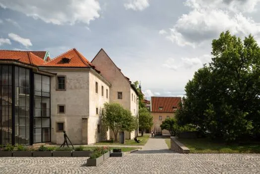 Historische stenen gebouwen met rode pannendaken en een moderne glazen uitbreiding in een binnenplaats met kasseien en groen