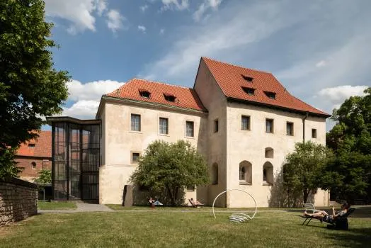 Een historisch gebouw met een rood pannendak en lichte stenen muren, omgeven door groen gras en bomen onder een blauwe hemel, met aan de linkerkant een moderne glazen uitbouw.