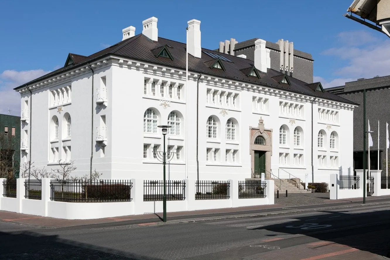 Edificio neoclásico blanco con ventanas arqueadas, techo oscuro y una valla de hierro a lo largo de la calle