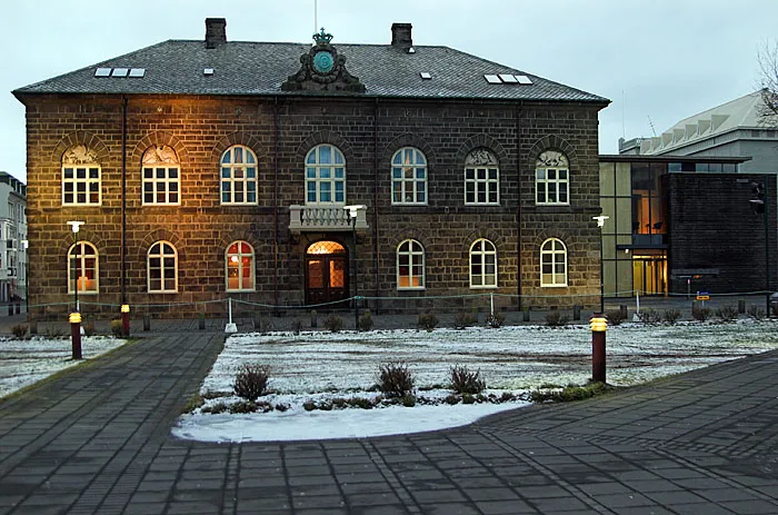 Un edificio histórico de piedra con ventanas arqueadas iluminadas desde el interior, ubicado frente a un patio cubierto de nieve al atardecer.