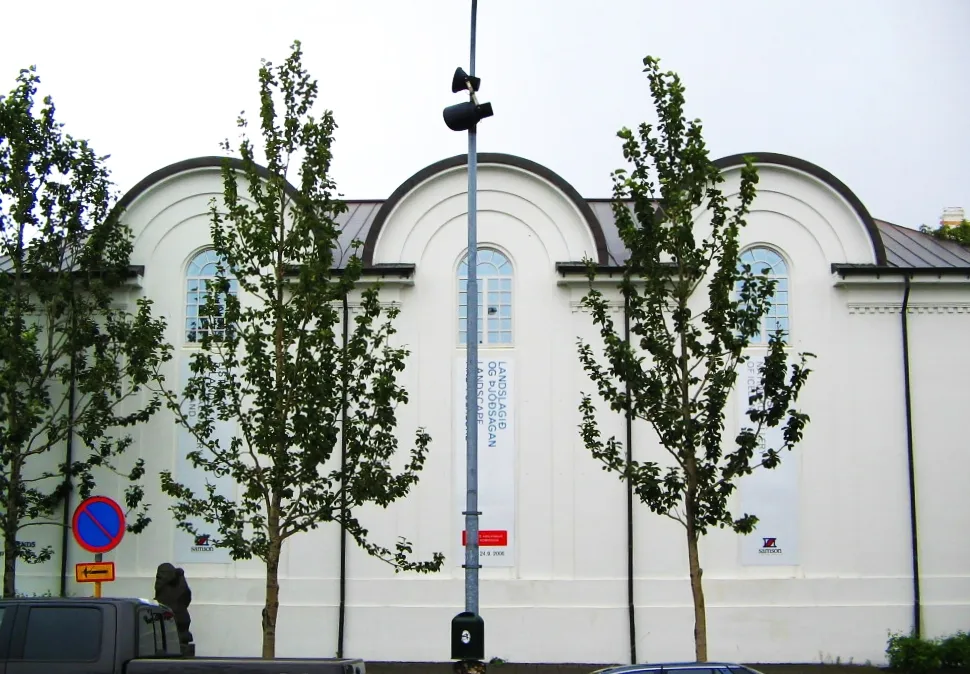 Fachada blanca con una serie de techos de arco redondeado y ventanas altas y estrechas con estandartes verticales, parcialmente oculta por árboles jóvenes y una farola central.