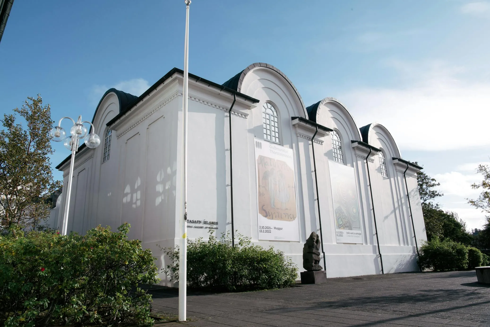 Edificio blanco con una línea de techo arqueada distintiva y grandes pancartas de exposición en la fachada, rodeado de vegetación y un asta de bandera bajo un cielo azul