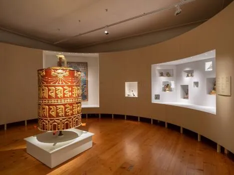 Een groot, kleurrijk gebedswiel tentoongesteld in een galerij met gebogen muren en verlichte vitrines