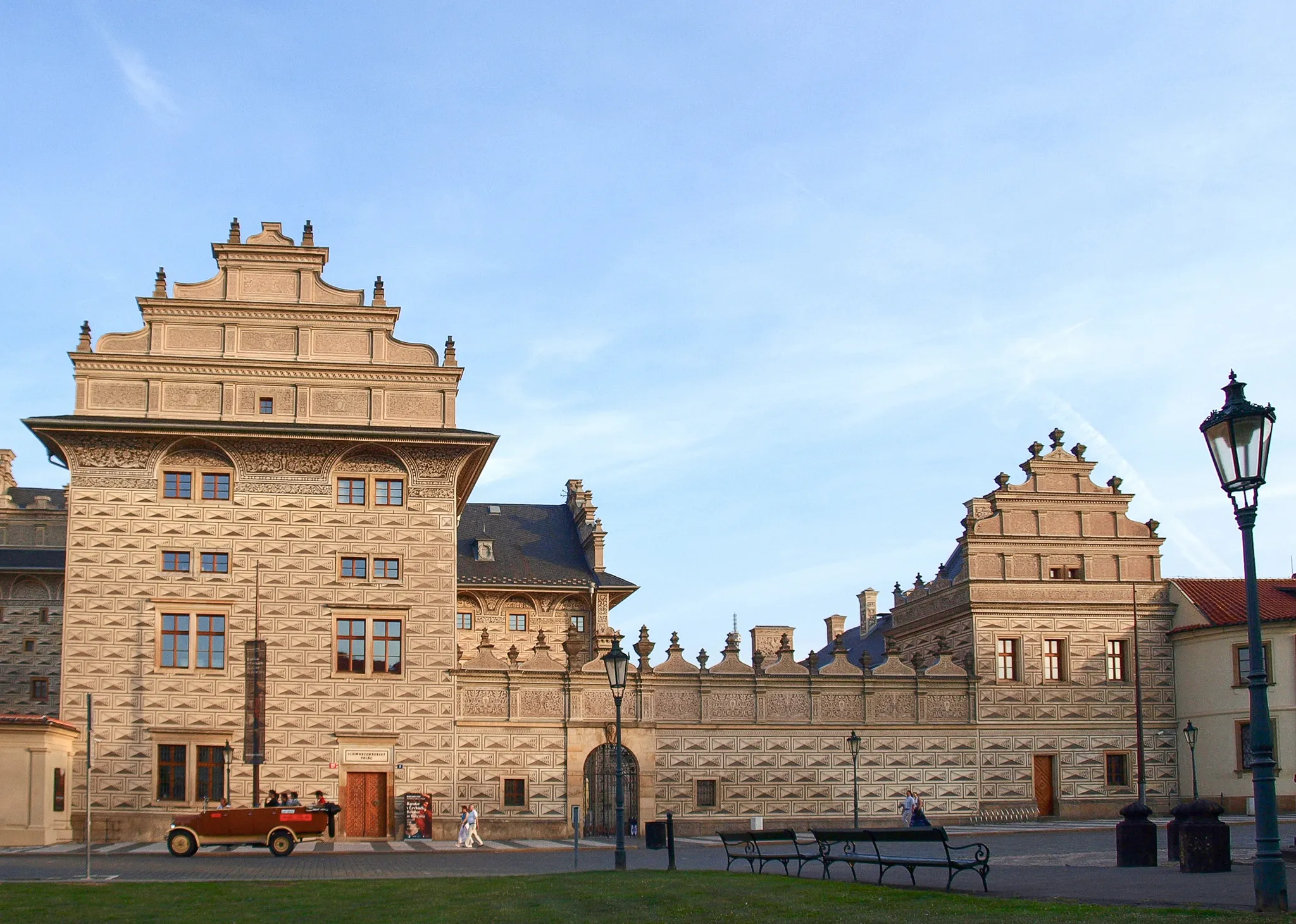 Pałac Schwarzenberg ze swoją architekturą renesansową, z attykami schodkowymi i kamienną fasadą, pod błękitnym niebem z zabytkowym samochodem i ludźmi na pierwszym planie.