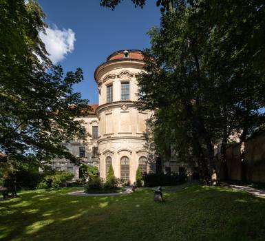 Barokowy pałac z zakrzywioną fasadą i kopułą, otoczony zielonymi drzewami i trawą pod błękitnym niebem