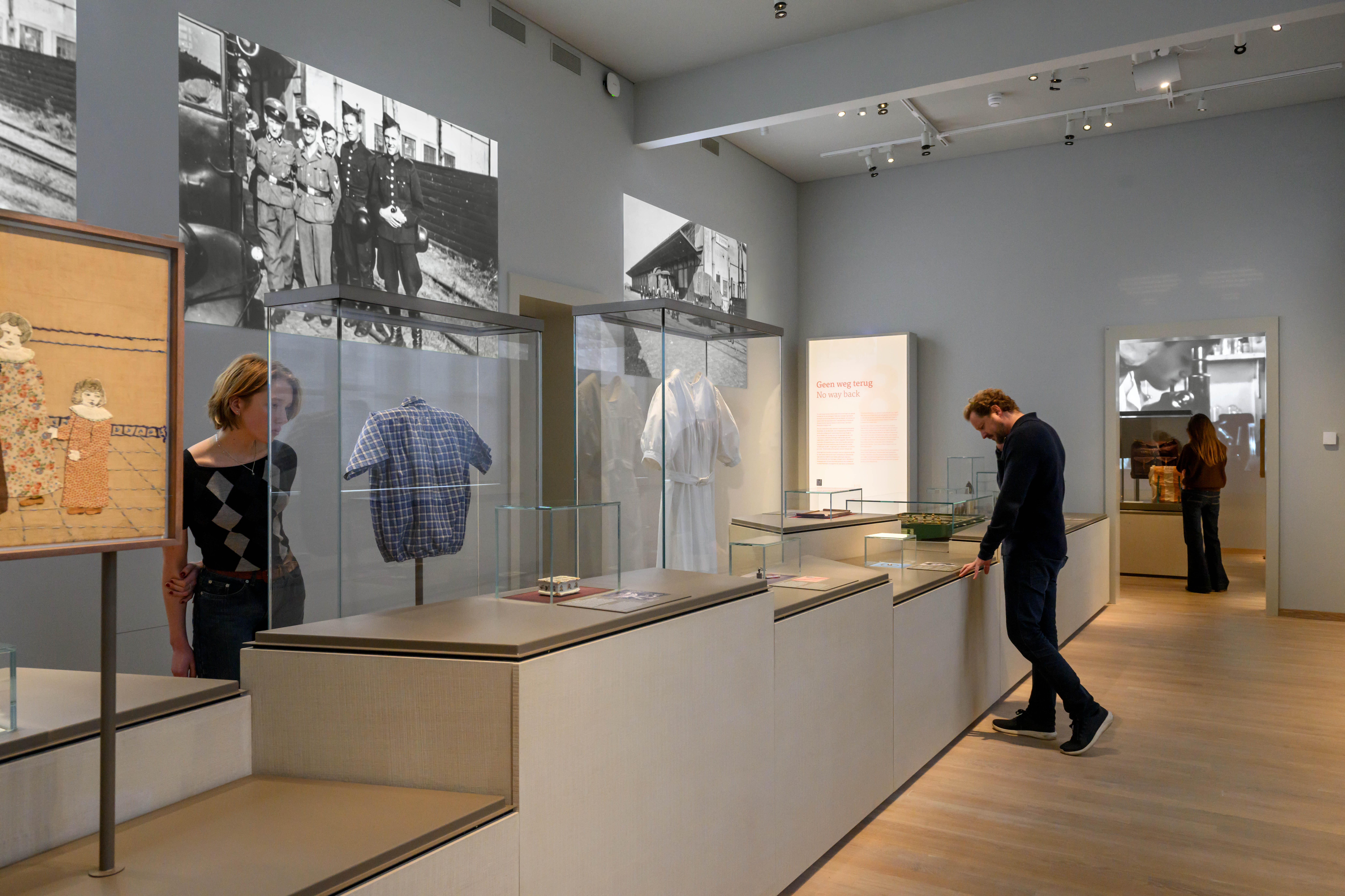 Przestrzeń wystawiennicza muzeum ze szklanymi gablotami zawierającymi artefakty odzieżowe, czarno-białe historyczne fotografie na ścianach oraz zwiedzających oglądających wystawy