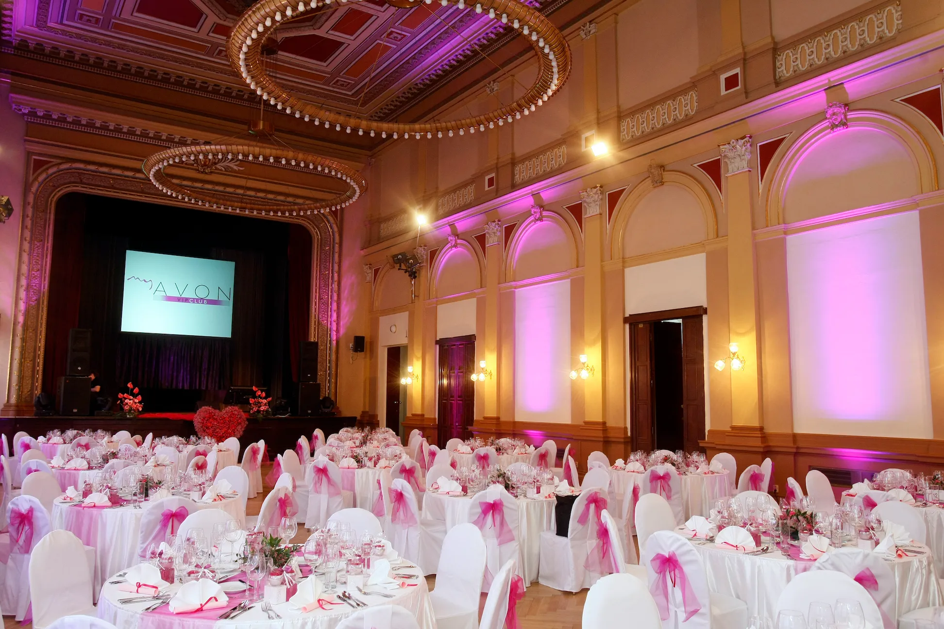 A grand hall with white draped tables and chairs, pink accents, and ornate ceilings with circular chandeliers and arched windows.
