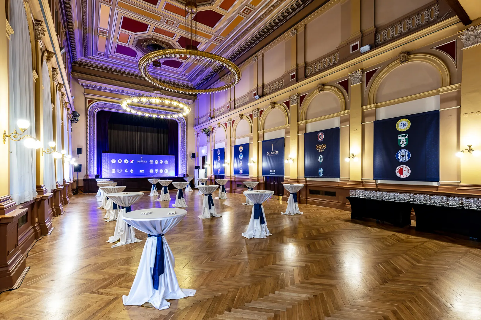 ornate hall with high ceiling, chandelier, wooden floor, and tables set up for an event