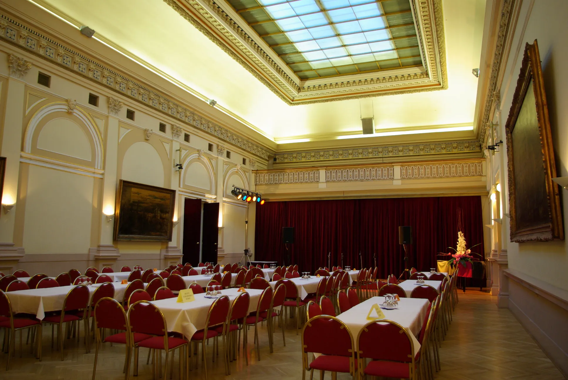 Interior of a grand hall with red chairs, white tablecloths, and a high ceiling with a skylight and decorative moldings